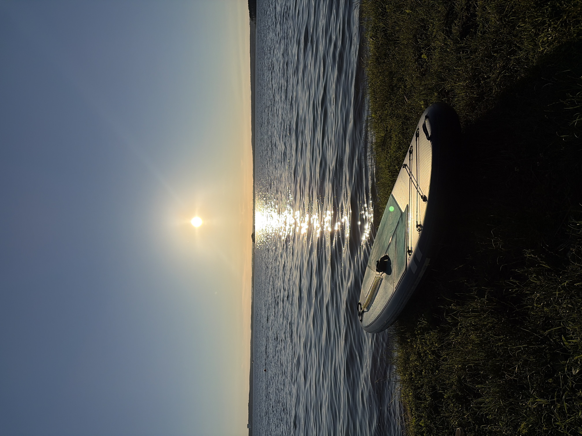 This is a landscape photograph taken at sunset, likely in the Ждановіцкі сельскі Савет region of Belarus. The sun is low on the horizon, casting a warm glow across the scene and creating shimmering reflections on the surface of a calm body of water, possibly a lake or river. The water is rippled, indicating a gentle breeze.
On the right side of the frame, a paddleboard is resting on the grassy bank of the water. The paddleboard is light-colored with a few subtle markings. It is angled towards the water, suggesting it is ready for use or has just been brought ashore. The grass appears lush and green, illuminated by the setting sun, with a long shadow cast by the paddleboard onto the darker, unlit grass further from the water.
The sky is a gradient of soft blue and pale yellow, typical of a sunset. The overall mood of the image is peaceful and serene, evoking a sense of tranquility and the end of a pleasant day spent outdoors. The time of day is clearly indicated by the low angle of the sun and the warm, long shadows. There are no visible people or text in the image.