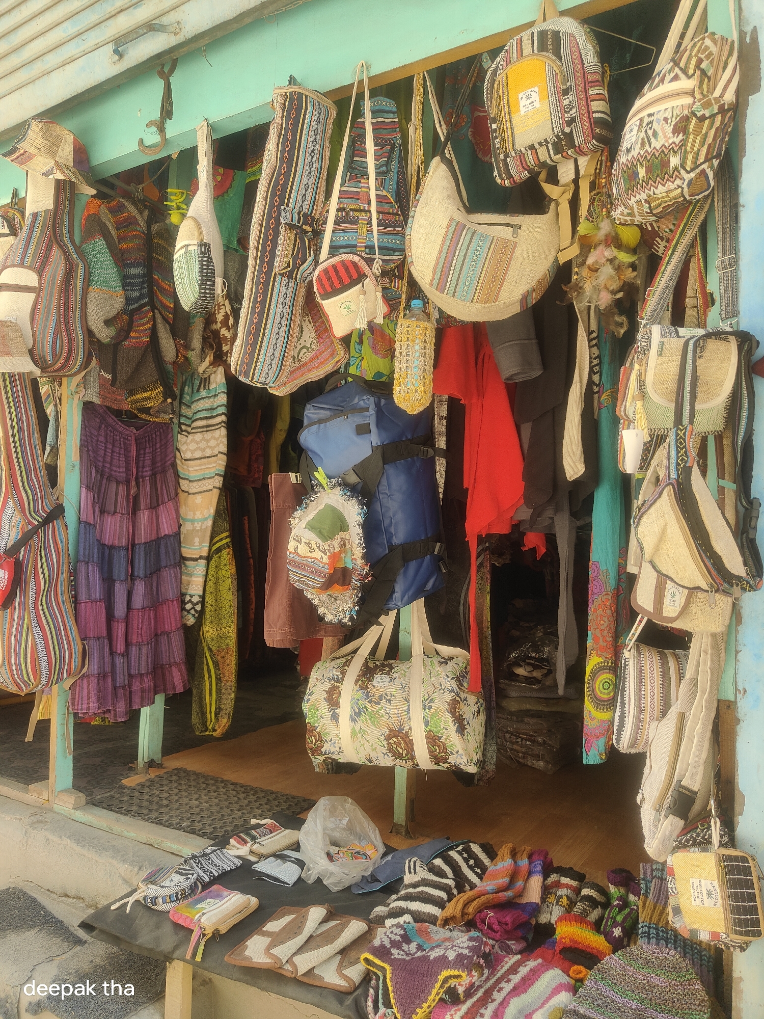 The image depicts a storefront in Leh, India, selling a variety of handcrafted bags, textiles, and accessories. The stall is set up outdoors, with merchandise displayed on hangers, hooks, and a table. The items for sale are colorful and feature intricate patterns, common in Tibetan and Ladakhi craftsmanship.
Specific items visible include:
* **Bags:** Numerous bags of various sizes and styles are hanging, including backpacks, shoulder bags, and duffel bags. Many are made from woven fabrics with geometric and ethnic designs. Some have leather or woven straps.
* **Textiles:** Several skirts and garments are displayed, also featuring bright colors and patterned fabrics. These appear to be made from lightweight materials like cotton or linen.
* **Accessories:** Smaller items such as wallets, pouches, and possibly hats or gloves are laid out on a table in the foreground. Some of these also showcase the characteristic colorful patterns. A few items are also displayed in a clear plastic bag.
* **Decorations:** Some of the bags have decorative elements like tassels or feathers.
The storefront itself has a teal-colored facade and appears to be constructed from wood. The background shows a dimly lit interior, suggesting the stall might be part of a larger market or shop. The ground is a mix of wooden flooring and a black mat. The overall impression is a vibrant display of local crafts, likely a common sight in tourist areas of Leh. The watermark "deepak tha" is visible in the bottom left corner of the image.