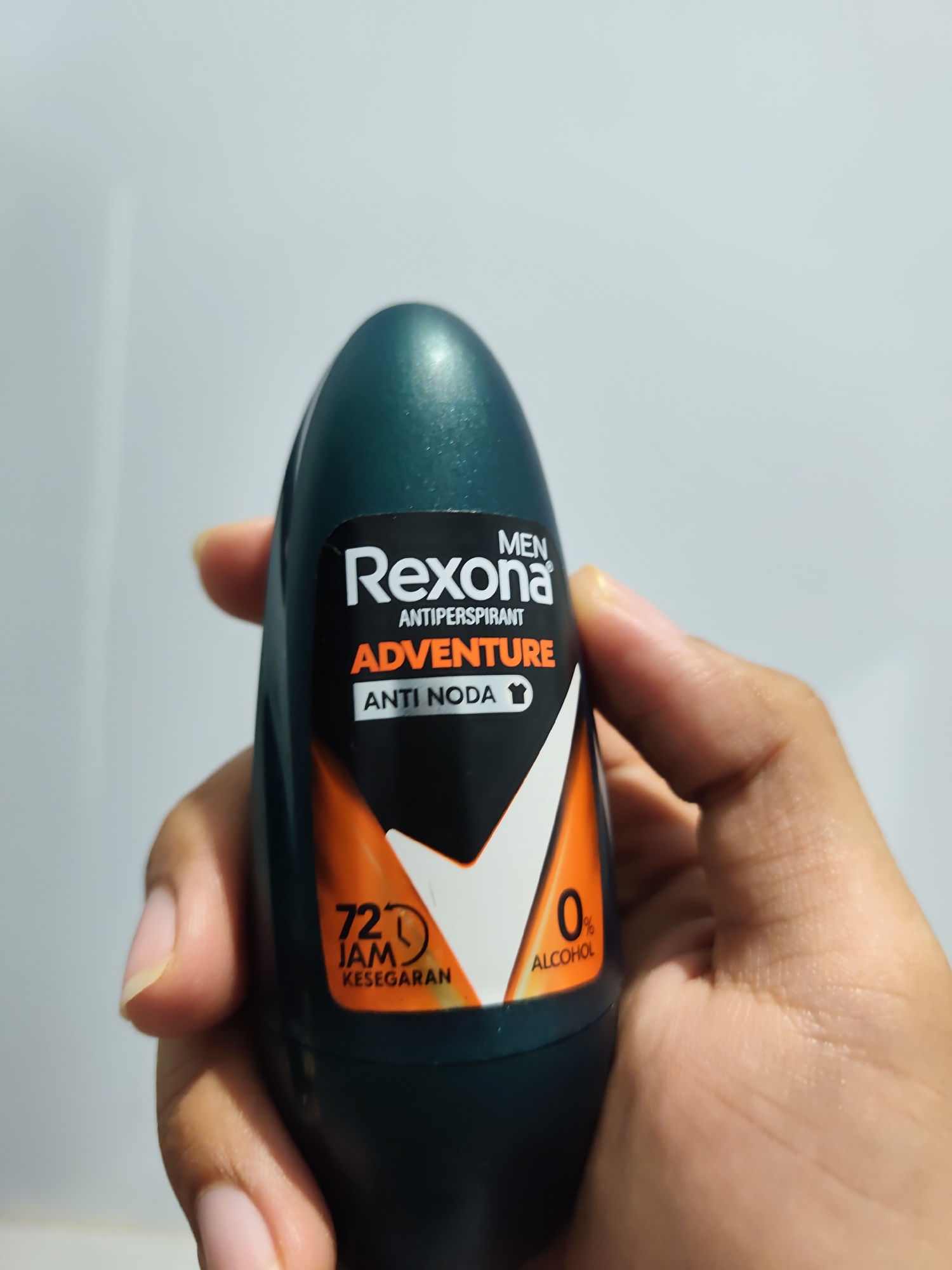 A person's hand holds a dark green roll-on antiperspirant bottle against a plain, light gray background. The bottle is oriented vertically, with the label facing the viewer. The label is predominantly black with orange and white text and graphics. Prominently displayed is the brand name "Rexona" in white. Below it, "ANTIPERSPIRANT" is in smaller white text. The word "ADVENTURE" is in large orange text, with "ANTI..." in white text and a small t-shirt icon to its right. To the left of the label's center, a graphic indicates "72 JAM KESEGARAN" (72 hours of freshness) in white text with a clock icon. To the right, "0% ALCOHOL" is in white text. The background appears to be a wall or a plain surface. The lighting is even, with no harsh shadows. The scene is indoors and the overall composition focuses on the product. The location of Pamotan, Indonesia, is not discernible from the image.