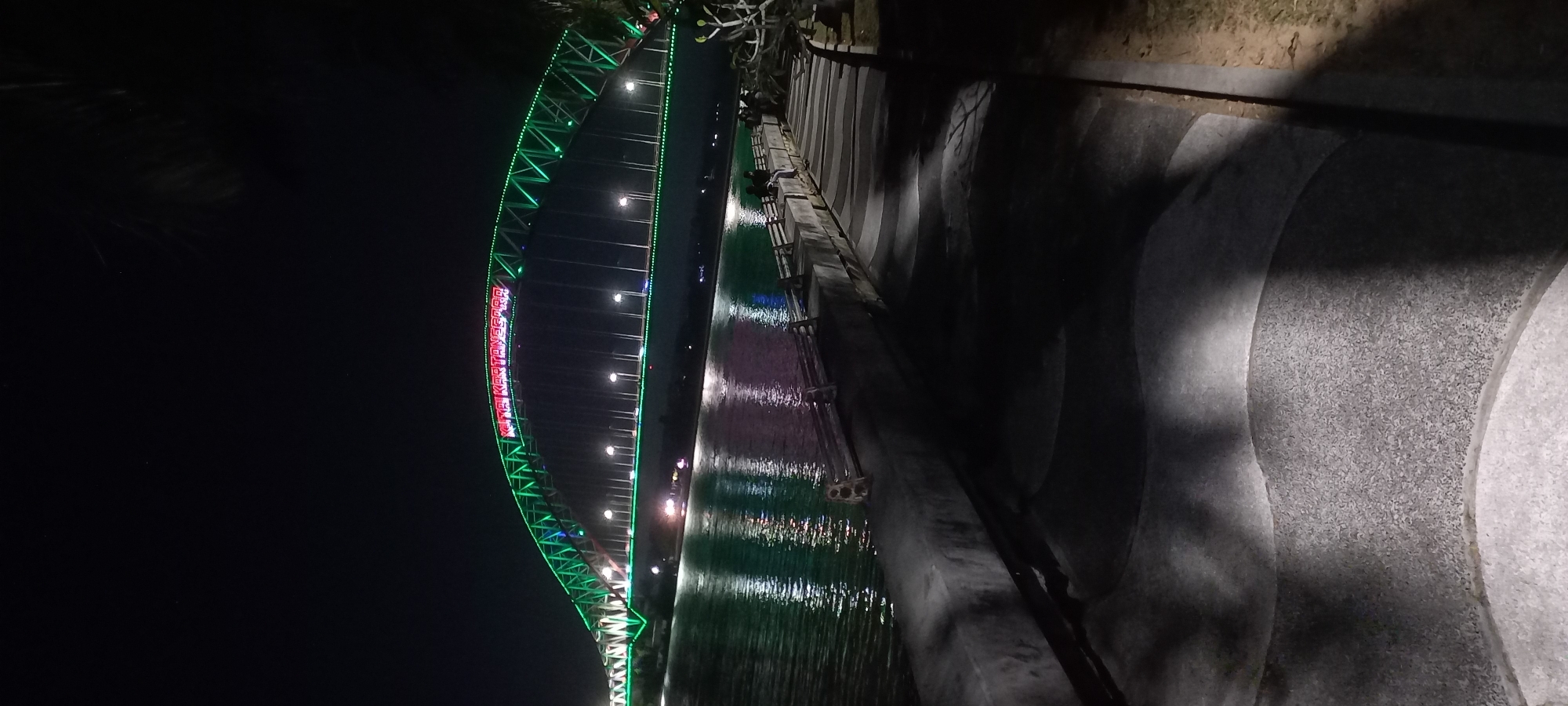 This is a nighttime, vertical shot of the Kartanegara Bridge in Teluk Dalam, Indonesia. The bridge is illuminated with bright green lights along its arch and supports, with red lights spelling out "KITA KARTANEGARA" across the apex of the arch. The bridge spans a body of water, likely a river or bay, which reflects the bridge's lights. In the foreground, there is a concrete embankment or walkway with textured paving and shadows cast by lighting not visible in the frame. A railing runs along the edge of the walkway, separating it from the water. Several people are visible sitting on the embankment. The overall scene depicts a well-lit urban infrastructure feature at night.