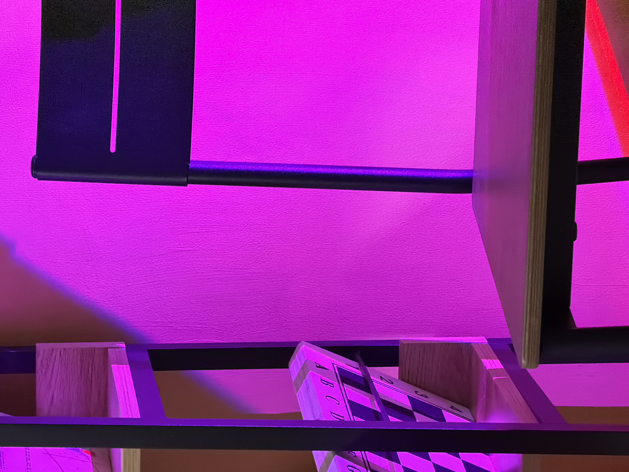 This image shows a close-up of a shelf unit, lit with purple and pink light. The shelves are made of light-colored wood with black metal frames. On one of the shelves, there is a board game with a checkerboard pattern, labeled with letters and numbers. The background is a flat surface, possibly a wall, illuminated with a solid, even pink light.
The location cannot be determined from the image content. It is impossible to confirm the location as Tel-Aviv, Israel, without additional information or context.