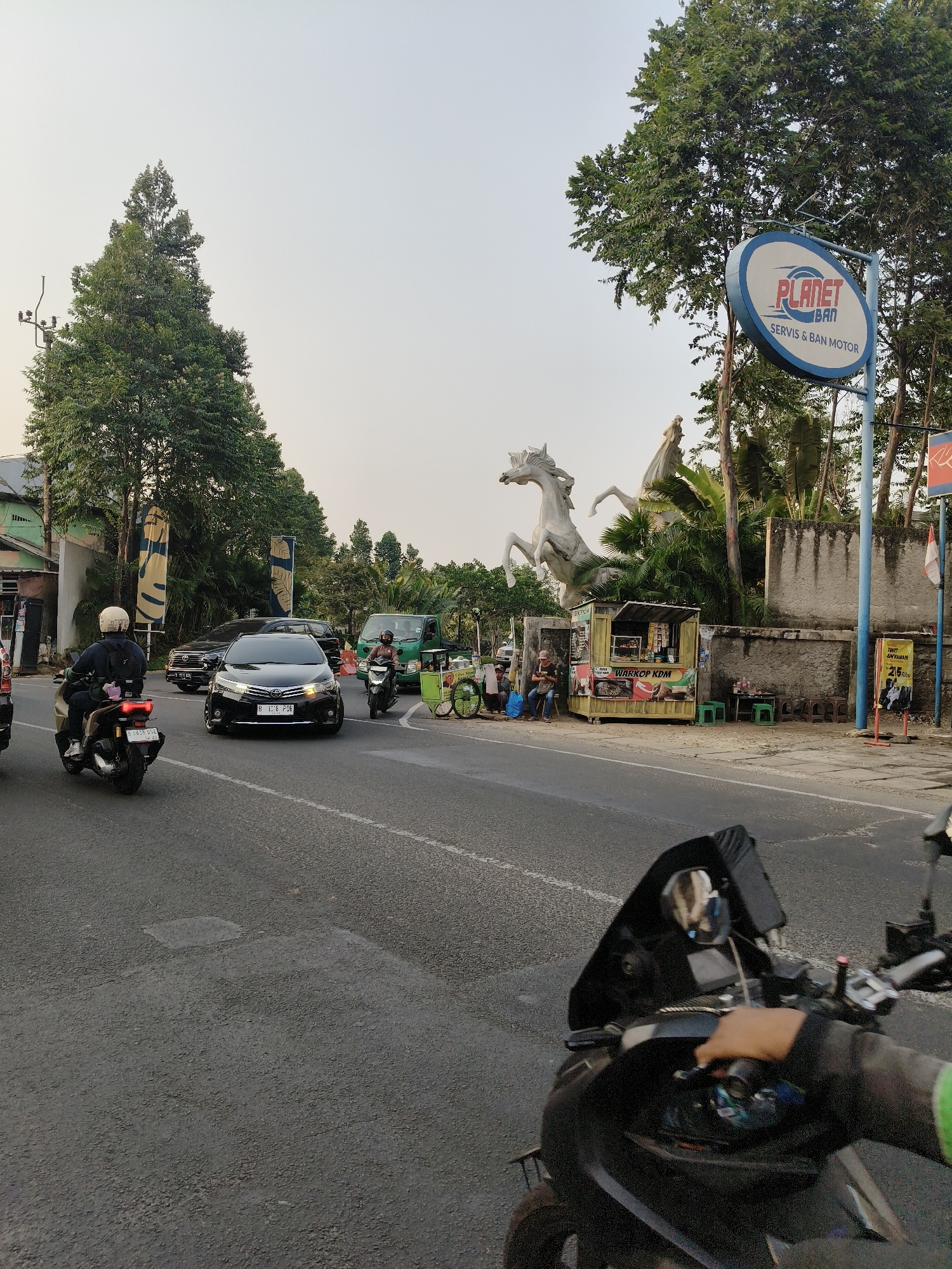 The image depicts a street scene in South Tangerang, Indonesia. In the foreground, a person is riding a motorcycle, their gloved hand on the handlebars. The motorcycle has a black body and a dark windshield. The street is paved and shows some wear.
Further down the road, a black car with license plate B 1318 PDG is visible, driving in the same direction. To the left of the car, another motorcycle with a rider wearing a white helmet is also on the road. In the background, a green truck is driving away.
On the right side of the street, there's a "Planet Ban" sign, indicating a tire service. Next to it, a small food stall labeled "Warkop KDM" is set up. Behind the stall, a large white sculpture of two horses rearing up is a prominent feature. Several people are seated around the food stall. Trees line the street, and a grey wall forms a backdrop behind the sculpture and stall. The sky is overcast.