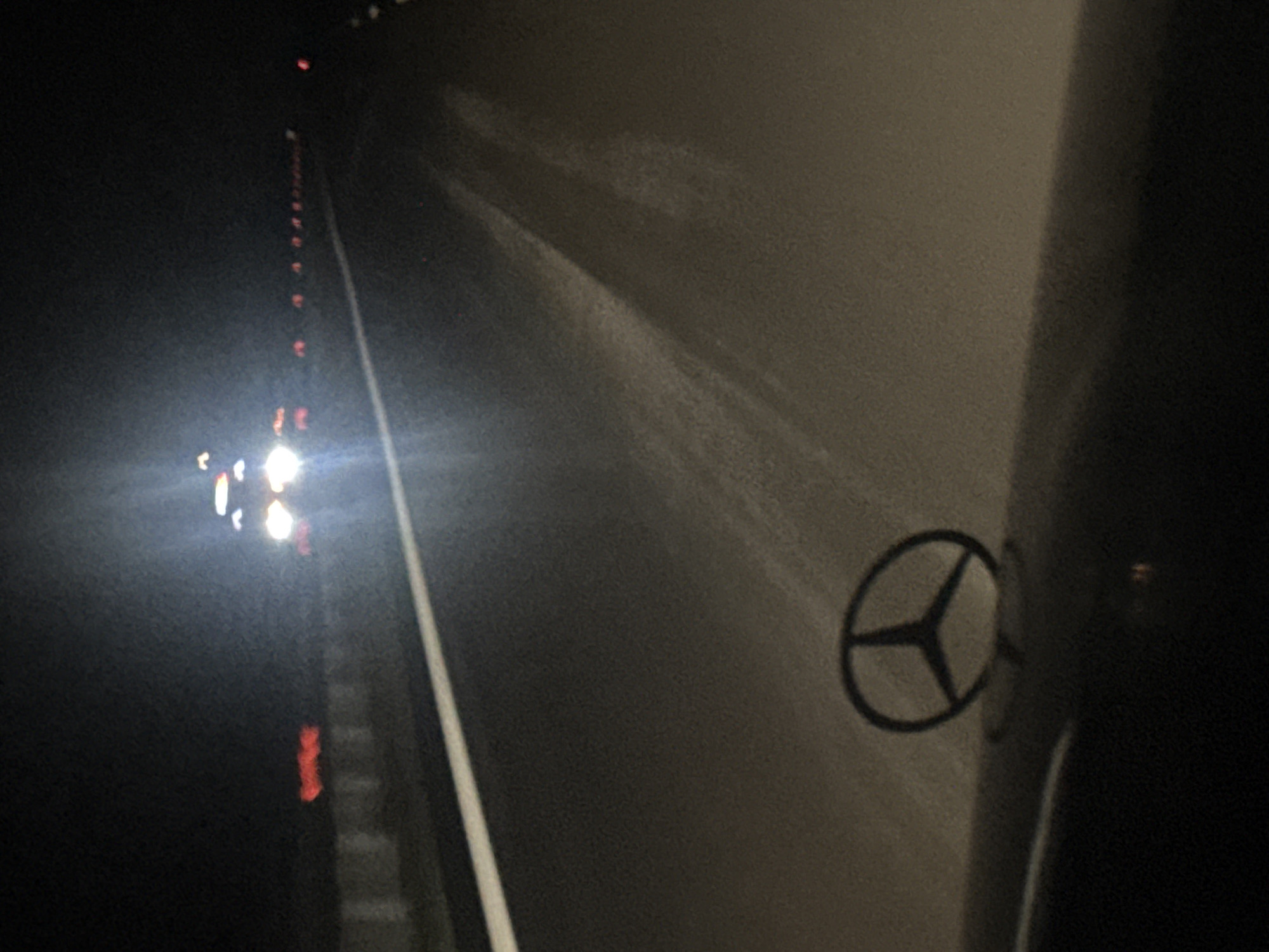 The image depicts a nighttime scene on a road, likely a highway, with a car's hood ornament, a Mercedes-Benz star, prominently in the foreground. The road has a white line marking its edge. In the distance, there are headlights of oncoming vehicles, some with red taillights visible, suggesting traffic. The overall lighting is dim, with the car's headlights creating bright glare. The image does not provide enough visual information to determine the specific location within the city of Zlatina, Bulgaria.