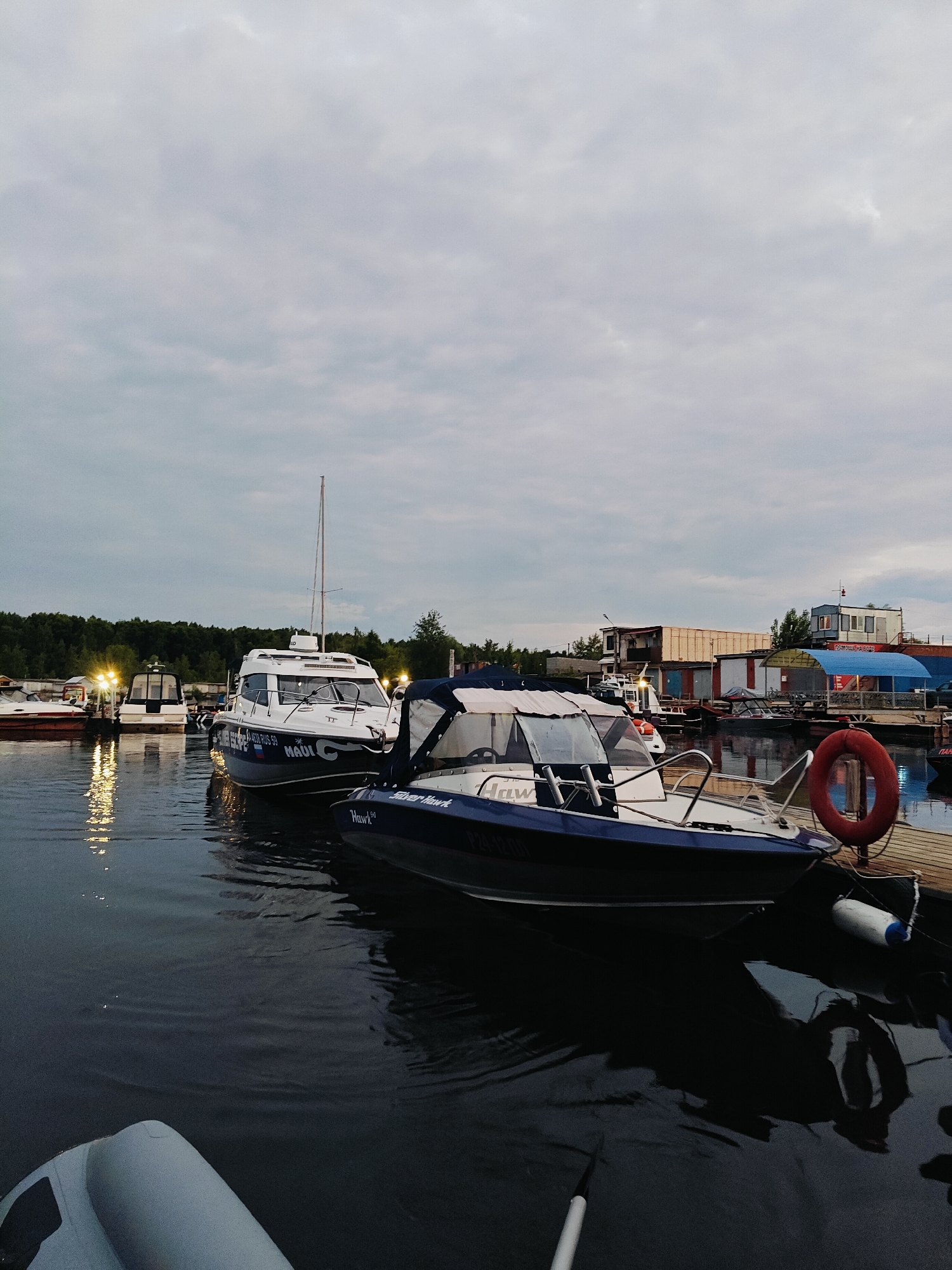 The image depicts a serene scene at a marina in Chaikovsky, Russia, likely during the early evening or twilight hours, given the diffused light and illuminated dock lights. The sky is overcast with a soft, cloudy texture.
Several boats are moored at the dock, reflecting on the calm, dark water. In the foreground, a sleek, dark blue and white motorboat with a covered canopy is prominently featured. The words "Silver Hawk" are visible on its side, along with a registration number that appears to be "7244 IZ 59". Another motorboat, white with blue accents, is docked next to it, displaying the name "MADI" and a registration number "4630 RU5 59". Further down the dock, a tall sailboat with a white mast stands out against the cloudy sky. Other smaller boats and structures, possibly storage units or small buildings, are visible in the background, lined with lights that cast a warm glow on the water. A red life preserver hangs on a post near the motorboat, and white and blue buoys are tied to the dock. The overall mood is peaceful and quiet, suggesting the end of a day of boating or a calm evening by the water. The setting is clearly a marina or a boat harbor on a body of water.
