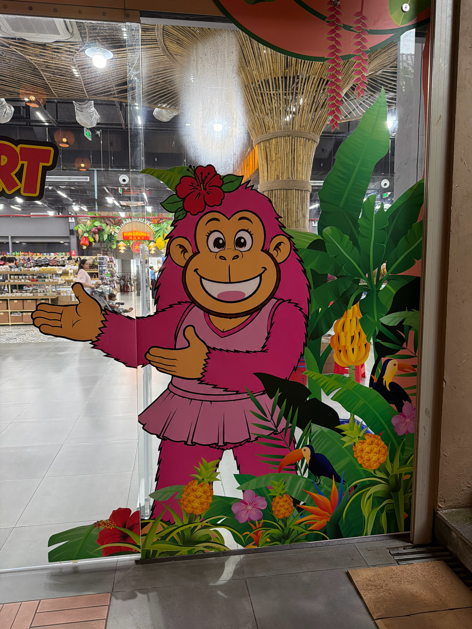 An exterior view of a shop entrance in Phu Quoc, Vietnam, features a large, cartoonish pink monkey decal on the glass door. The monkey is depicted with a cheerful expression, wearing a pink dress and a red flower in its hair. It is surrounded by a tropical-themed mural with large green leaves, pineapples, and colorful flowers. In the background, through the glass, shelves stocked with various goods and shoppers can be seen inside the store, suggesting a retail environment. The floor outside the shop is tiled, with a brown mat placed near the door. The overall scene depicts a welcoming and playful storefront.