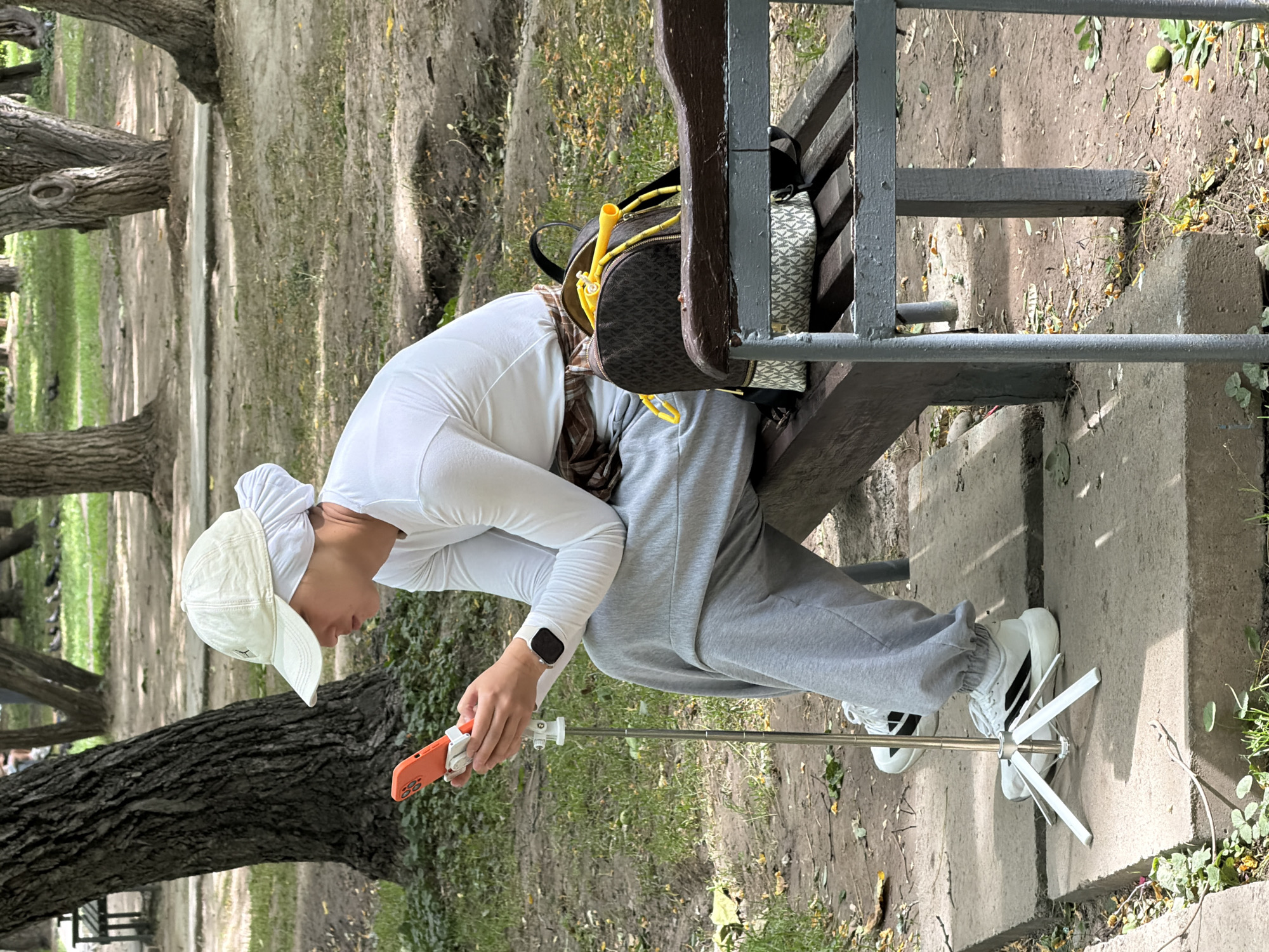 A person wearing a white cap, white long-sleeved shirt, and grey sweatpants is sitting on a park bench and looking at an orange smartphone. The person is holding the phone with both hands, and the phone is attached to a selfie stick with a tripod base. The person is wearing white sneakers with black accents. A brown and tan patterned backpack with yellow rope accessories is on the bench next to the person. The background consists of trees, grass, and dirt pathways, suggesting an outdoor park setting. The lighting indicates it is daytime.