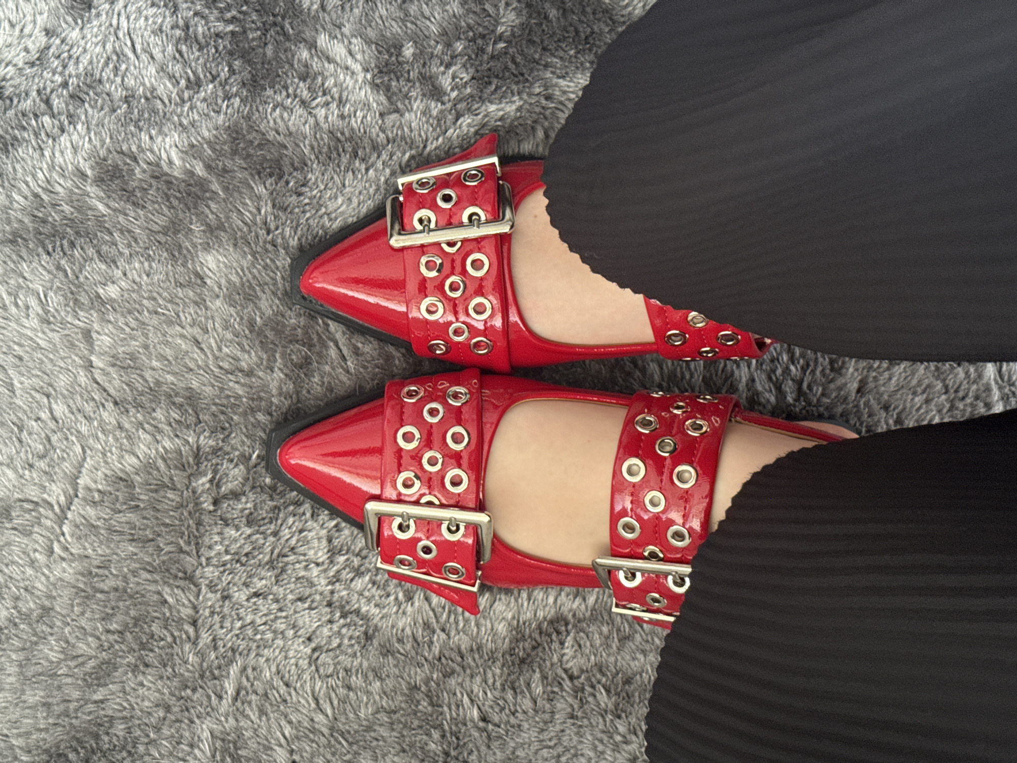 A top-down view captures a person's feet adorned in bright red patent leather shoes, resting on a textured gray shag rug. The shoes are pointed-toe flats, each featuring two wide straps with silver-colored buckles and multiple small silver eyelets covering the straps. The patent leather surface reflects light, adding to the vibrancy of the red.
The person is wearing black pants that partially obscure the top and outer sides of the shoes. The pants appear to have a subtle pleated or ribbed texture, particularly noticeable on the right side of the frame. The gray rug, which serves as the background, is thick and shaggy, with visible individual fibers creating a soft, uneven surface.
The image focuses on the footwear and its immediate surroundings, presenting a fashion-oriented shot. There is no discernible activity beyond the static placement of the feet. No specific time of day, weather, or emotions are evident, and no text is visible in the frame. The scene is set in Mexico City, Mexico.