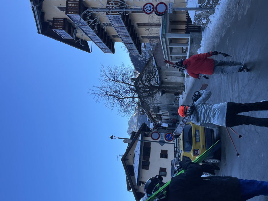 The image depicts a vibrant winter street scene in Courmayeur, Italy, under a clear, bright blue sky, suggesting daytime. Several individuals, likely skiers or snowboarders, are seen from behind or the side, walking along a street partially covered in snow and ice. They are dressed in winter attire, including jackets (red, light grey, dark) and helmets (orange, black), and are carrying skis with poles or snowboards.
The scene features traditional alpine architecture with multi-story buildings lining the street. Balconies on the buildings and the bare branches of a prominent tree in the middle ground are visibly frosted, indicating cold temperatures. In the background, snow-capped mountains are discernible. A yellow car, possibly a Fiat, with the Italian license plate "EZ 765YW" is parked on the side of the road.
Visible street signs include a circular white and red sign indicating a height restriction of "2.70 m," another circular red and white prohibitory sign, and a rectangular white sign with Italian text "eccetto residenti e proprietari" (except residents and owners). The ground exhibits patches of ice and snow reflecting the bright sky, contributing to the crisp, winter atmosphere. The activity suggests movement through a ski resort town, possibly to or from the slopes.