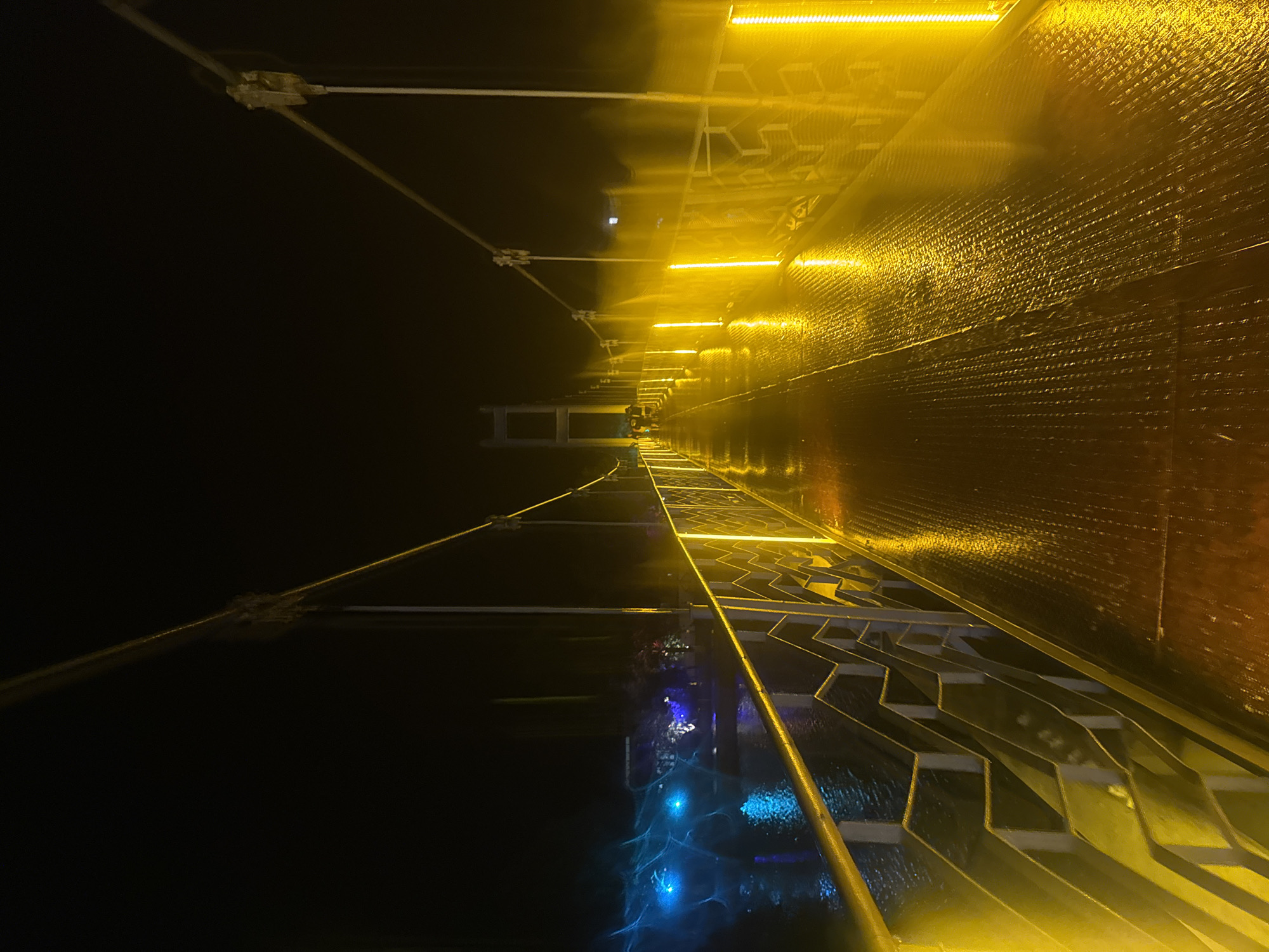 The image depicts a suspension bridge at night in Poti, Georgia. The bridge deck is illuminated by yellow lights, and the surface appears wet, reflecting the light. The railing along the side of the bridge is also lit with yellow lights, creating a zig-zag pattern. In the distance, a larger bridge structure can be seen. On the left side, the water below the bridge is illuminated by blue lights, and there are some trees and buildings visible along the waterfront, also with colored lights. A few people are walking on the bridge in the distance. The sky is dark, indicating it is nighttime.