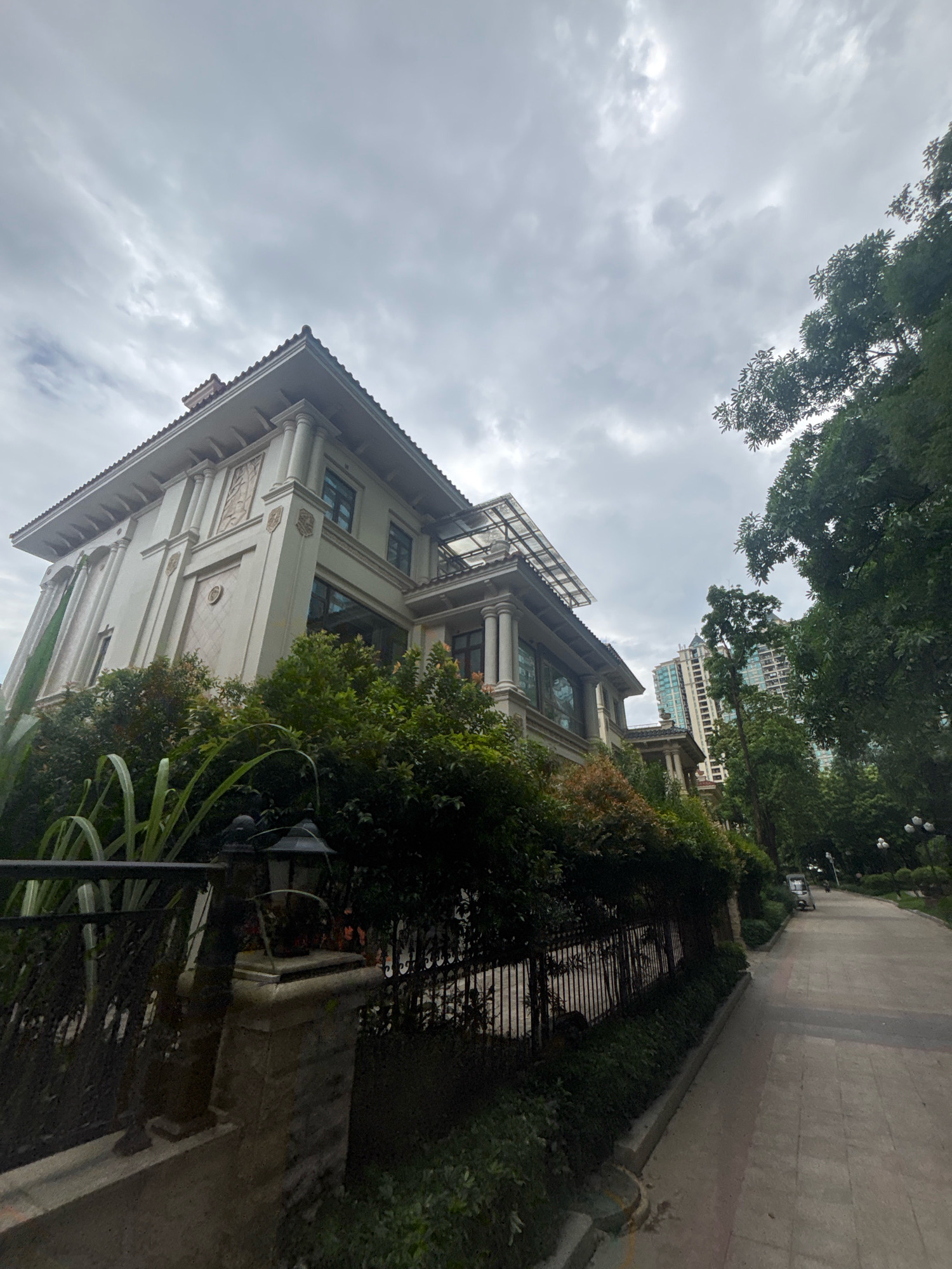 The image shows a wide-angle, low-angle view of a large, multi-story residence in the city of Nanhai District, China. The house is light-colored with a tiled roof, classical columns, and decorative relief work on its facade. It is surrounded by lush green trees and manicured bushes. A black metal fence with intricate detailing runs along the front of the property, with a stone pillar supporting a vintage-style lamp. To the right of the residence, a paved walkway curves away into the distance, bordered by more trees and vegetation. In the background, a tall, modern skyscraper can be seen peeking above the trees, suggesting an urban setting. The sky is overcast with grey clouds. No people are visible in the image.
