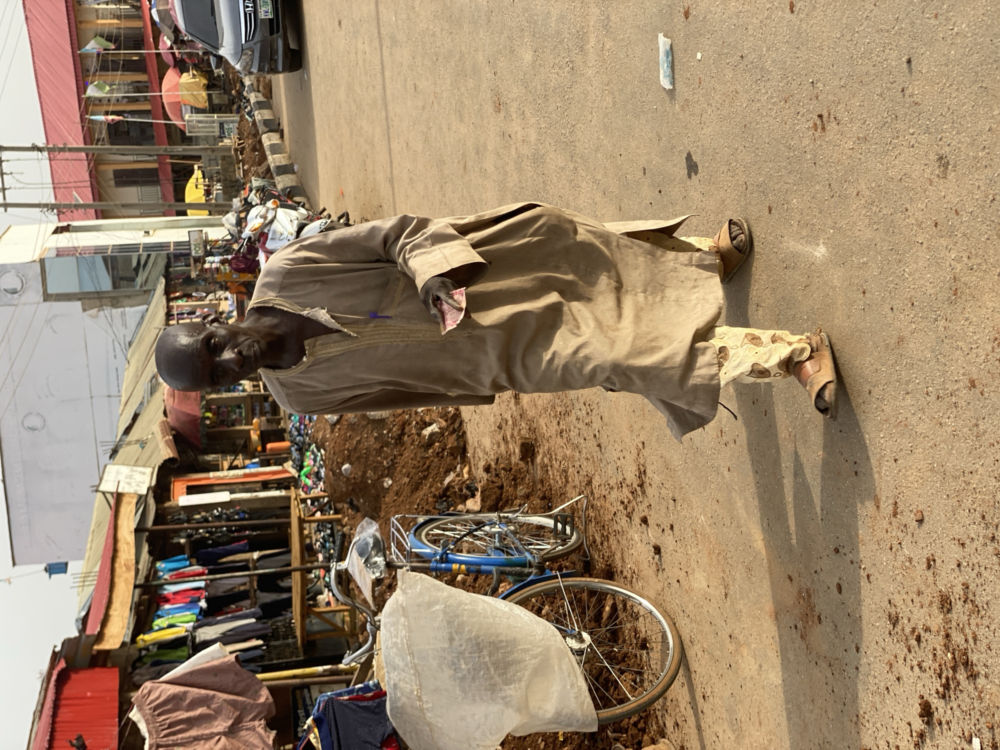The image captures a vibrant street scene in what appears to be a market area in Kafanchan B, Nigeria, under bright daylight with strong shadows indicating a sunny sky. The central subject is a dark-skinned man, partially bald, standing with a calm expression, looking towards the viewer. He wears a long, loose-fitting tan robe (likely an agbada or similar traditional garment) over patterned light-colored trousers and brown sandals. In his left hand, he holds several pieces of paper currency.
To his right, a blue bicycle with a white plastic bag covering its front section leans against a pile of brown dirt and debris. In the background, numerous market stalls are visible, displaying various garments like shirts and trousers on wooden racks, suggesting active commerce. Buildings with reddish roofs and light-colored walls line the street, while utility poles and wires crisscross the sky. A black car, possibly a Honda, is parked further down the street, with part of its license plate visible showing "KML 340...". Several motorbikes or scooters are also parked nearby. A sign with the text "POS" (Point of Sale) can be seen in the distance. The ground is an asphalt surface, interspersed with patches of dirt and debris.