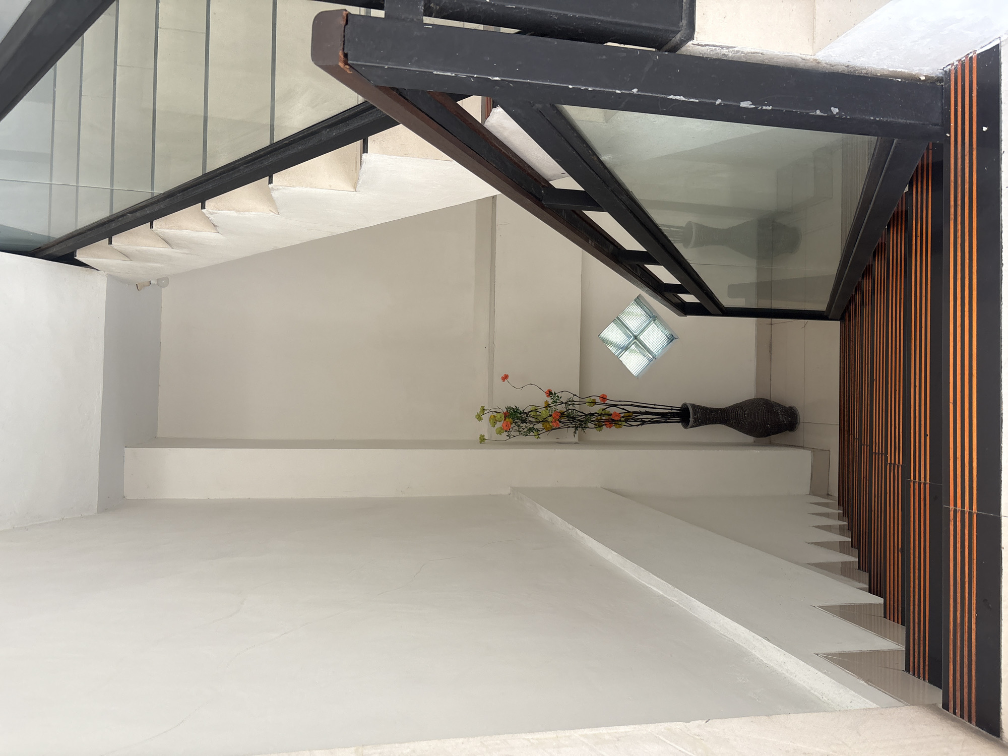 An interior shot looking down a staircase. The staircase has black treads with orange stripes and white risers. A dark gray vase with artificial flowers is placed on the floor next to the staircase. The staircase has a black metal railing with glass panels. The walls are painted white. A small square window made of glass blocks is visible on the wall. The scene is lit by overhead lighting.