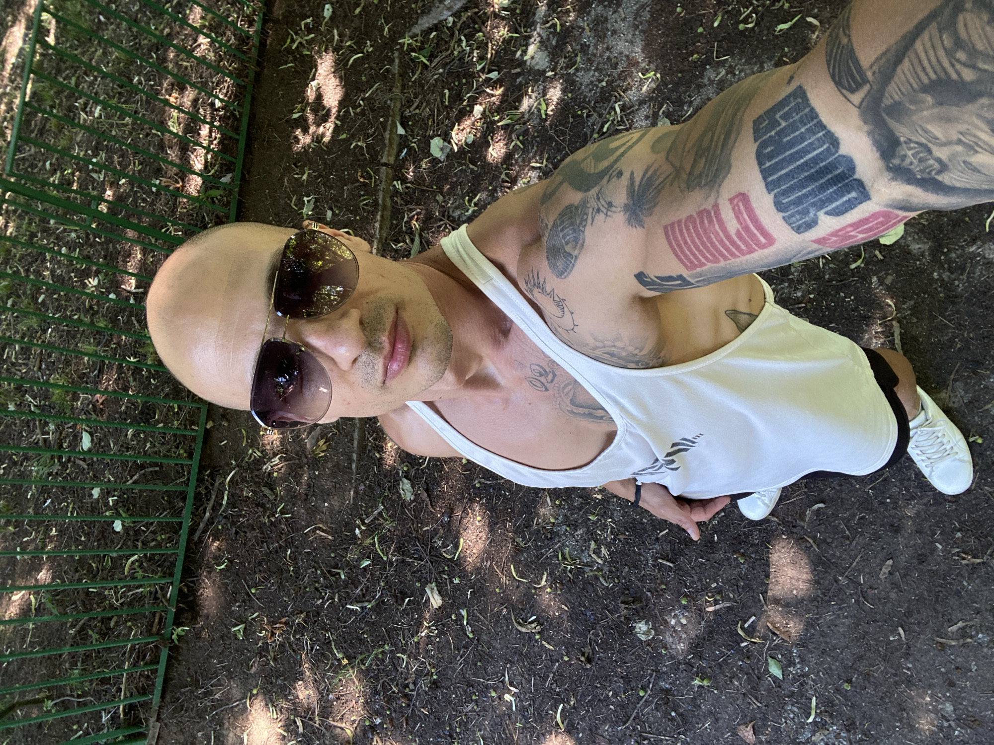 A bald man wearing sunglasses and a white tank top is taking a selfie outdoors. He has tattoos on his left arm and chest. He is wearing white sneakers and black shorts. The background consists of dirt, leaves, and a green metal fence. The lighting suggests it is daytime and sunny, with dappled shadows on the ground. The location is identified as the city of Lovech, Bulgaria.
