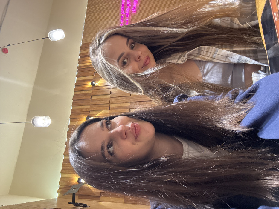 Two young women pose for a selfie indoors. One has long, light brown hair with blonde highlights, wearing a light grey tank top and a tan plaid shirt. The other has long, dark brown hair and wears a white top and a dark blue jacket. They are positioned close together, looking up at the camera. Behind them, there is a wooden wall with several small lights and a light beige wall with lights.