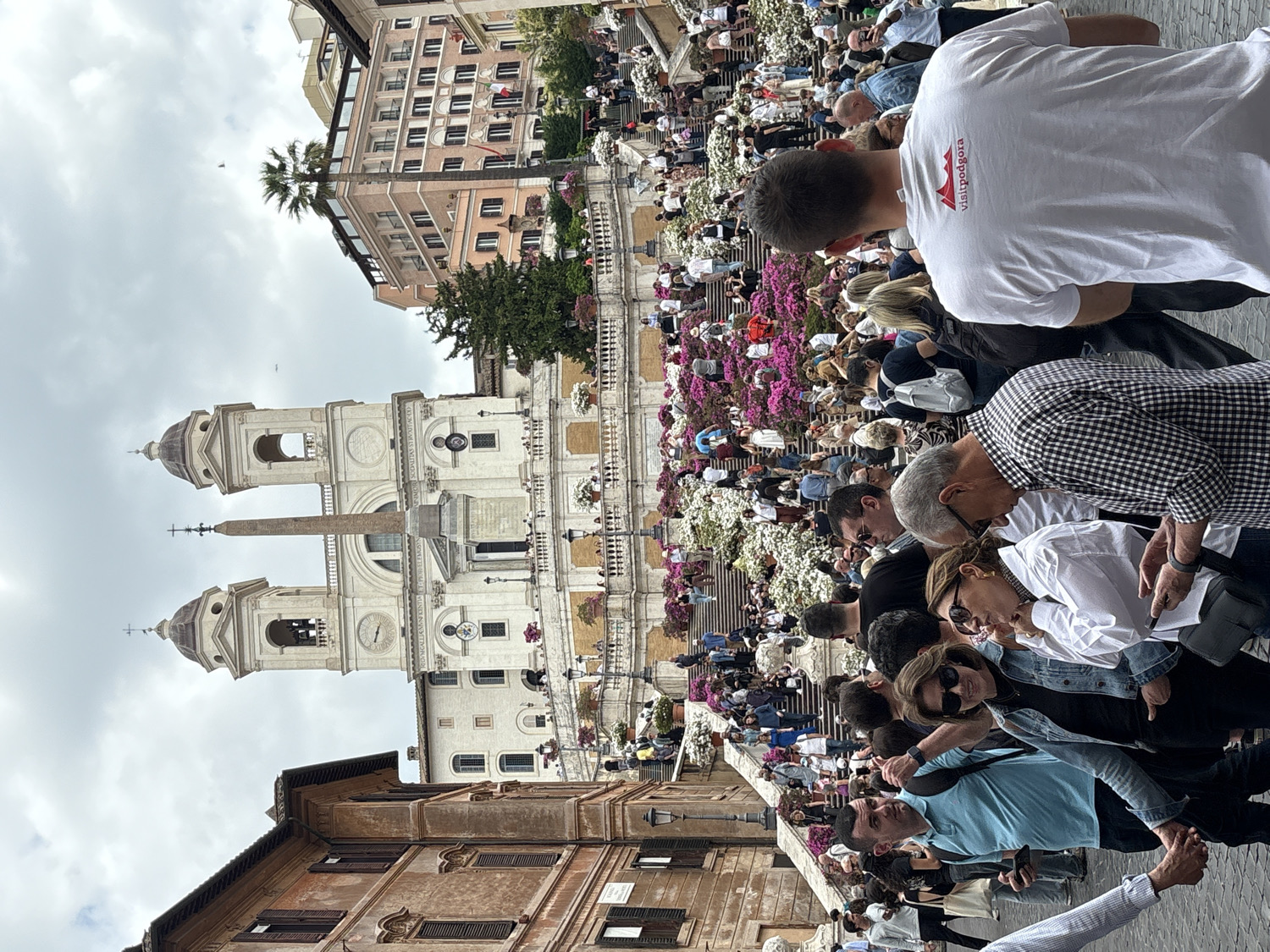 The image, rotated 90 degrees clockwise, captures a lively daytime scene at the iconic Spanish Steps (Scala di Trinità dei Monti) in Rome, Italy. A large crowd of people of diverse ages fills the wide stone staircase, some ascending or descending, others gathered on the steps, observing their surroundings. Many individuals appear to be tourists, engaged in sightseeing and socializing.
At the top of the steps stands the prominent Trinità dei Monti church, characterized by its twin bell towers and a distinct obelisk positioned in front. The church's facade features architectural details and what appears to be a Latin inscription, though it is not clearly legible from this distance. Historical buildings with multiple windows line the sides of the square, including a building with a palm tree visible in the upper left.
The steps are beautifully adorned with numerous flowering plants, predominantly vibrant pink and white azaleas, suggesting spring or early summer. The sky is overcast, indicating a cloudy but bright day.
In the foreground, among the crowd, a man wearing a white t-shirt is visible from behind. His shirt clearly displays the text "VISITPODGORA" alongside a stylized mountain or crest logo. The overall atmosphere is one of bustling urban activity at a popular landmark.