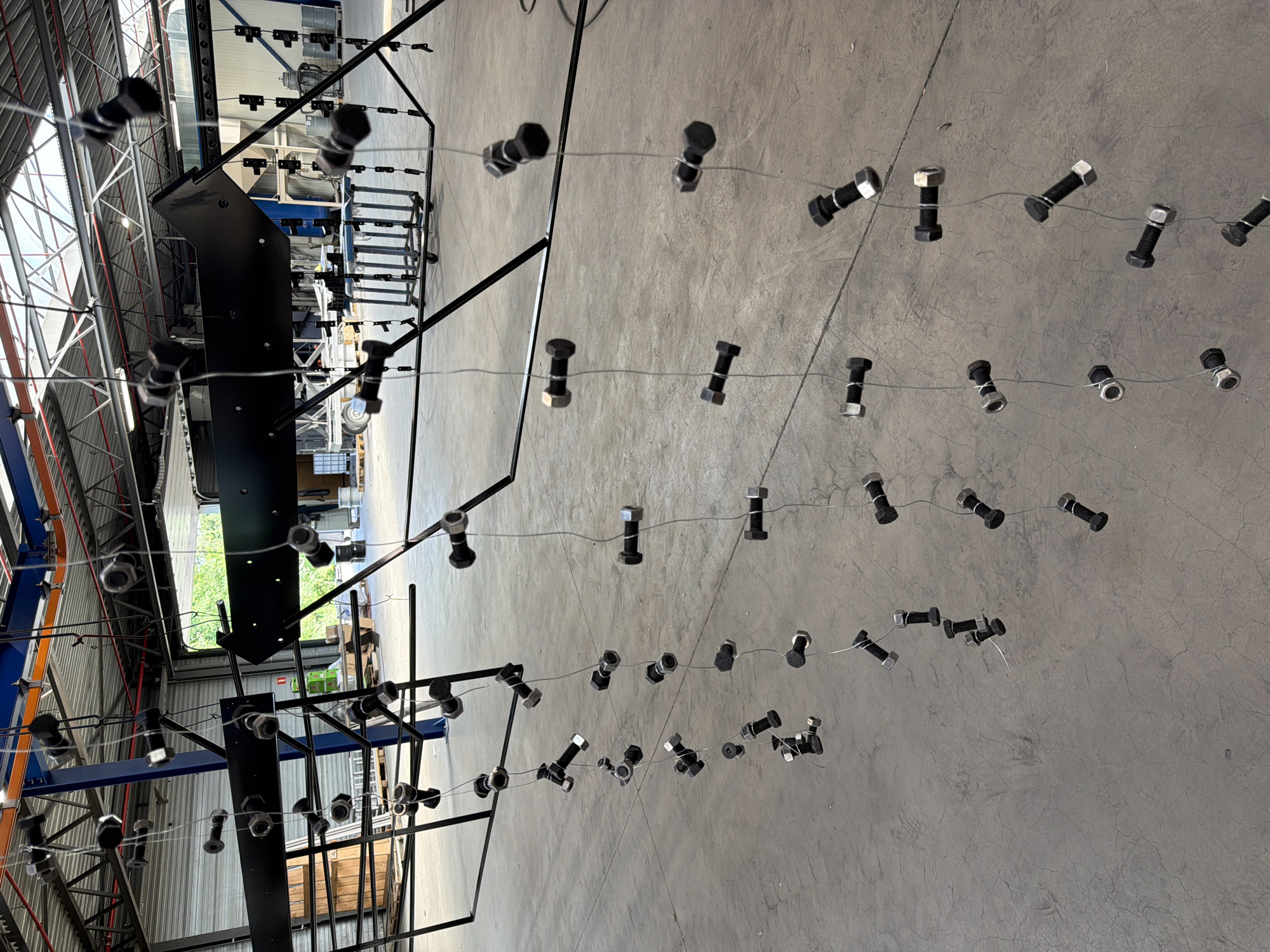 The image depicts an industrial setting, likely a workshop or manufacturing facility. Hanging from a black metal structure are numerous bolts and nuts, suspended by thin wires. These fasteners are arranged in vertical lines, creating a visually striking pattern. In the background, the facility's interior is visible, with industrial machinery, shelving units, and large overhead doors. The floor is a polished concrete surface. The presence of the bolts and nuts suggests they are either awaiting processing, such as painting or assembly, or are part of a decorative or artistic installation within the industrial space. The image was taken in Mol, Belgium.