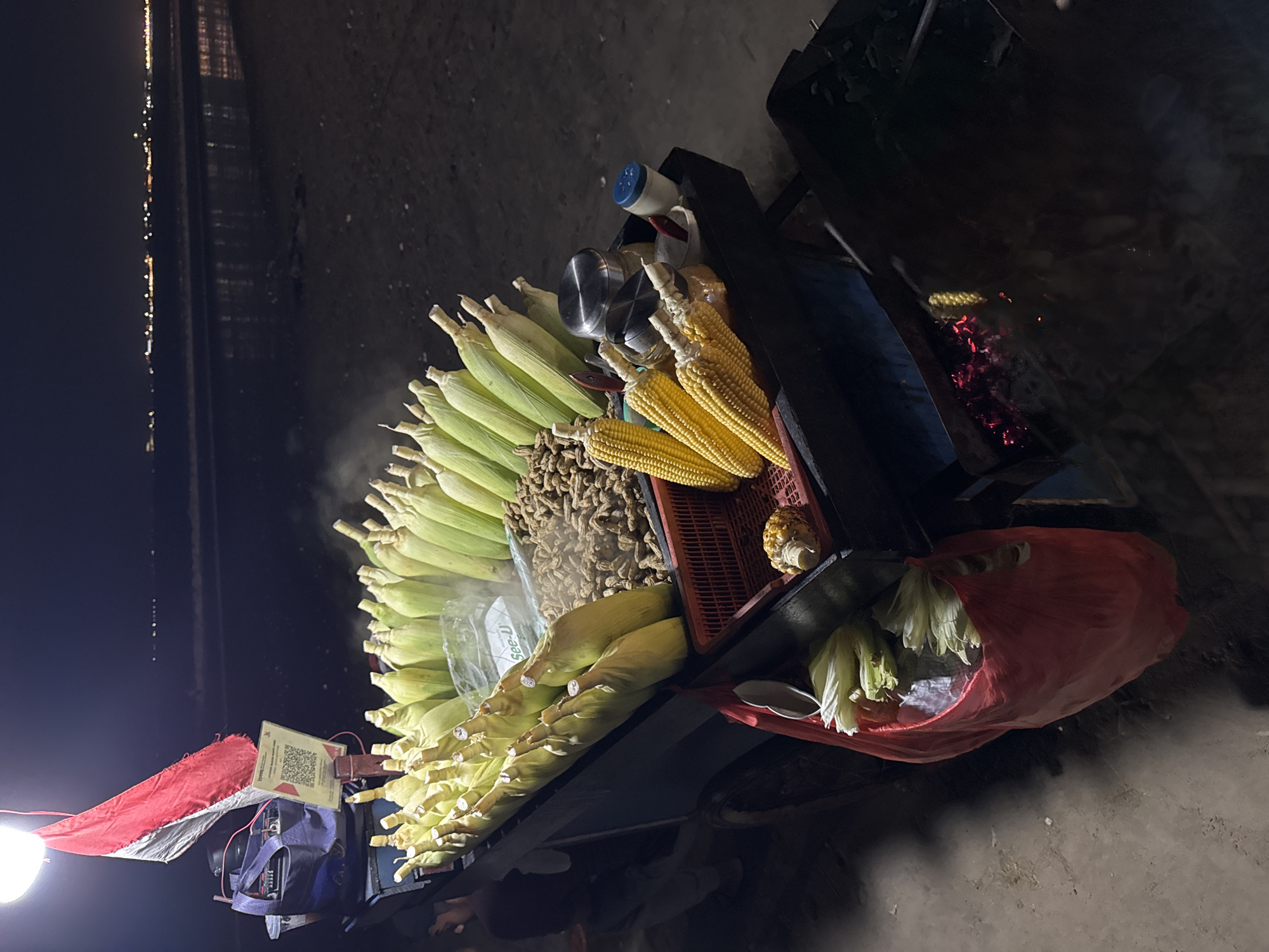 The image displays a street food cart operating at night, likely in Kuta Selatan, Indonesia, given the context and visible Indonesian flag. The cart is illuminated by a bare lightbulb on the left, casting a bright glow on the merchandise.
The primary items for sale are ears of corn, arranged in large piles. Many ears are still in their green husks, while others are shucked, revealing bright yellow kernels. A faint wisp of steam rises from some of the corn, suggesting it's being cooked or kept warm. Adjacent to the corn is a container filled with what appears to be peanuts or small nuts.
Towards the right side of the cart, a section reveals glowing red embers, indicating a heat source, likely used for roasting or heating the corn. Various containers, including metal shakers and jars, are neatly arranged on the cart. A red plastic bag on the lower right contains additional corn or husks.
In the dark background, a line of distant, scattered lights stretches across the horizon, possibly from a city, buildings, or along a coastline. The ground beneath the cart appears to be sandy or unpaved. A small red and white Indonesian flag is attached to the cart. For transactions, a stand displaying a QR code is visible, and the text "See-U" is readable on a plastic bag. No clear human figures are visible, but the setup implies a vendor is present.