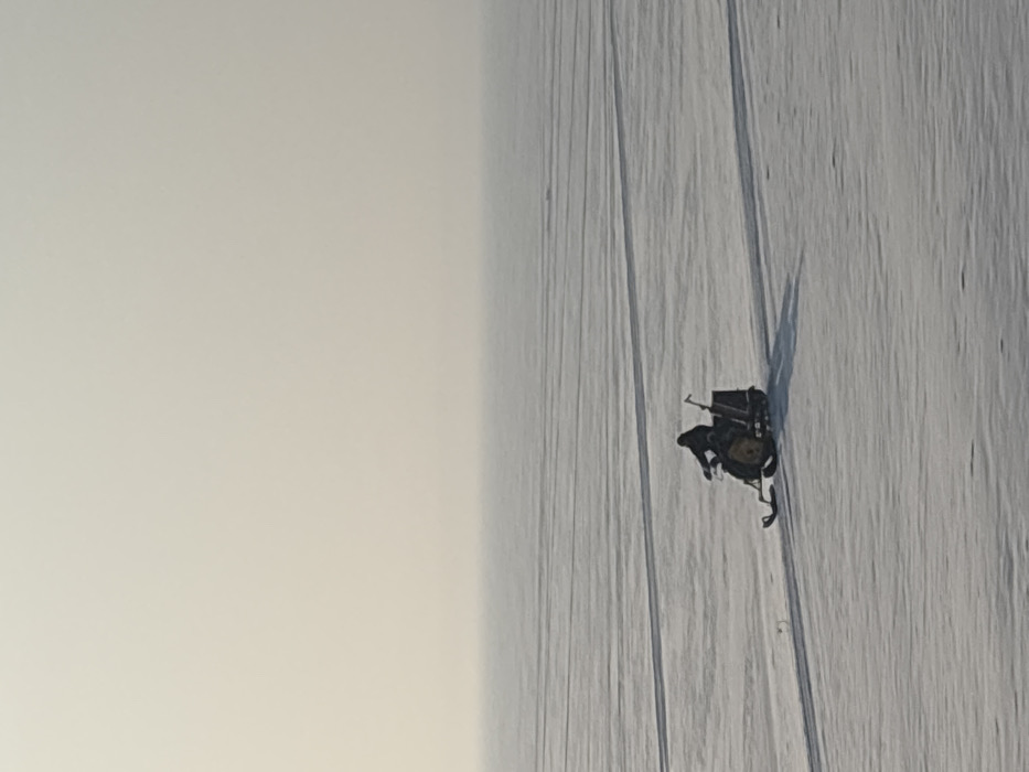 A single individual on a snowmobile traverses a vast, flat expanse of snow-covered ice, consistent with the frozen Kama Reservoir near Chaikovsky, Russia. The person, dressed in dark winter attire, operates a dark-colored snowmobile featuring yellow accents, moving from left to right across the frame. A dark, pole-like object, resembling an ice auger or shovel, is vertically secured to the rear of the snowmobile. The vehicle casts a distinct long shadow to its lower right on the snow, which also displays faint, parallel lines indicating prior passage. The environment is dominated by a uniformly pale, hazy sky that blends into the distant horizon, lacking any discernible features.