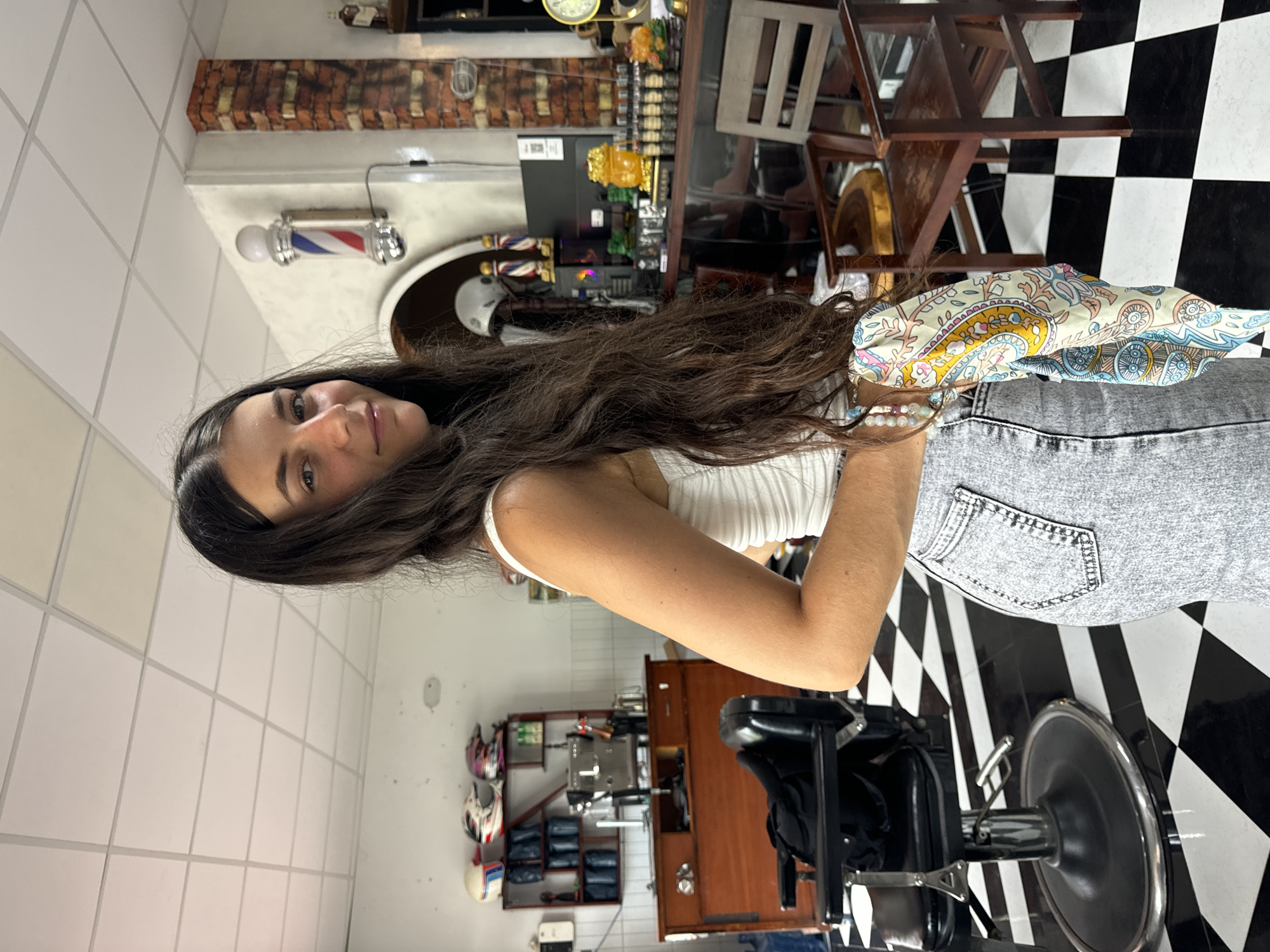 A young woman with long, dark hair stands in what appears to be a barbershop in Phan Thiết, Vietnam. She is wearing a white tank top and light-wash denim jeans. She holds a colorful scarf in her right hand. The barbershop has a distinctive black and white checkered tile floor, a vintage barber chair, and shelves filled with various items, including helmets, bottles, and what looks like a coffee machine. A classic barber pole is visible on a wall adorned with faux brick. Sunlight streams in, illuminating the scene and casting shadows on the floor.