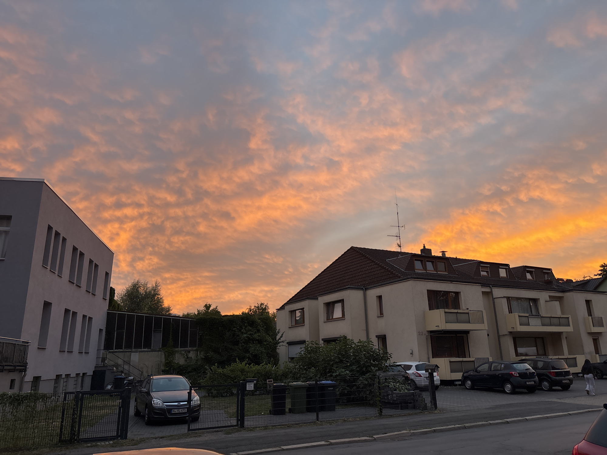 The image depicts a cloudy sky illuminated by the warm hues of a sunset, casting an orange and pink glow across the scene. In the foreground, a street runs along the bottom of the frame, with a sidewalk adjacent to parked cars. To the left, a modern, light-colored building with multiple windows is visible, partially obscured by a fence. Adjacent to this building is a darker, more enclosed structure with vertical windows. To the right, a multi-story building with a reddish-brown roof and balconies is situated. Several cars are parked in front of this building, and a person is walking away from the viewer on the sidewalk. Green trash bins are located behind a fence in front of the buildings. Trees and foliage are present, particularly behind the fence and between the buildings, suggesting a suburban or residential setting within a city. The sky is a prominent feature, with a delicate pattern of clouds contributing to the atmospheric quality of the image.