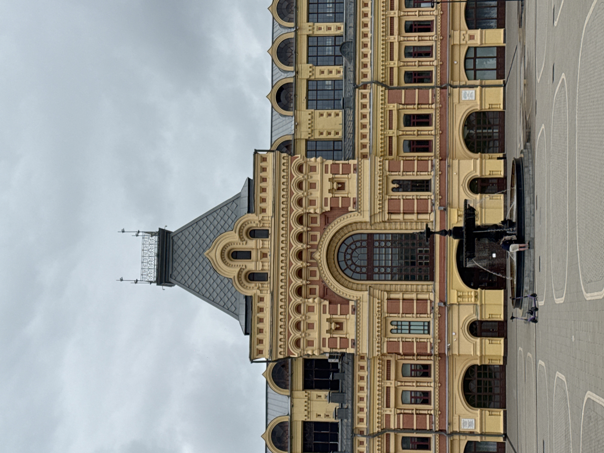 The image shows the Nizhny Novgorod State Academic Opera and Ballet Theatre, a prominent landmark in Nizhny Novgorod, Russia. The theater's ornate yellow and red facade, characterized by intricate architectural details and arched windows, is the central focus. A fountain is situated in the foreground, with water spraying upwards. A person is partially visible standing near the fountain. Two electric scooters are parked on the paved plaza in front of the building. The sky is overcast, contributing to the overall muted lighting of the scene. The plaza is paved with grey interlocking stones, with decorative curved patterns.