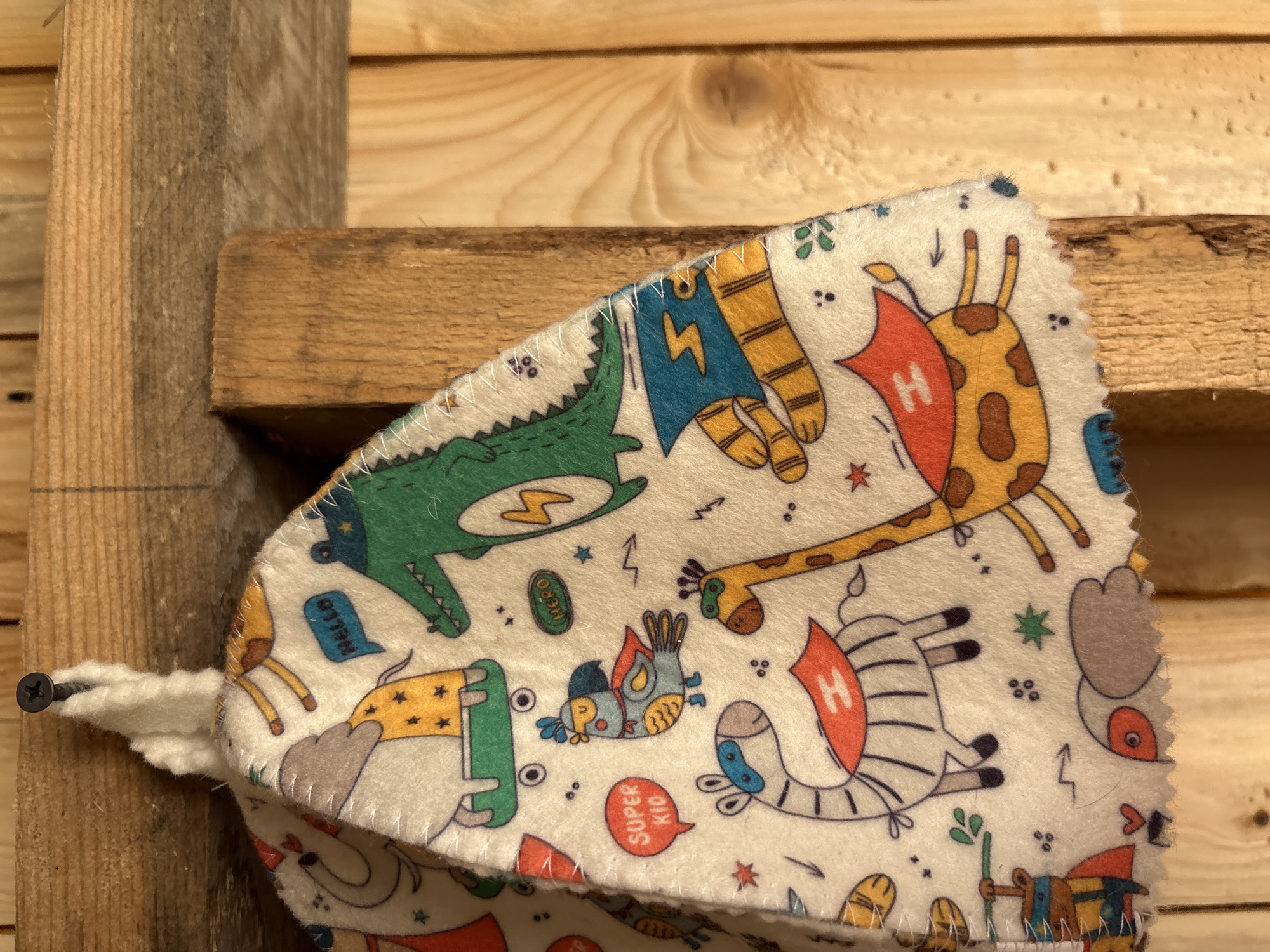 A white, fuzzy fabric item, possibly a cap or hat, decorated with colorful cartoon animal designs, is depicted hanging from a black Phillips-head screw. The screw is driven into a light brown, horizontally oriented wooden plank located in the upper portion of the frame. The fabric displays various anthropomorphic animals, many portrayed with superhero characteristics such as capes and masks. Prominent figures include a green alligator with a yellow lightning bolt emblem on its chest and a "HELLO" speech bubble, a zebra wearing a blue mask and a red cape with a white 'H', and a giraffe also featuring a green mask and a red cape with a white 'H'. A bird with a blue mask and a red cape is adjacent to a "SUPER KIO" speech bubble, with another "HERO" text bubble nearby. Partially visible are an elephant on a green skateboard and a tiger with a blue cape and a lightning bolt. The edges of the fabric item are finished with visible white stitching. The background primarily consists of multiple light to medium brown wooden planks, exhibiting distinct wood grain and texture consistent with unfinished lumber. A vertical wooden plank is visible on the right side of the hanging fabric item. No individuals are present in the image. The visual information does not provide evidence to confirm a specific geographical location, such as the city of Italmas, Russia.
