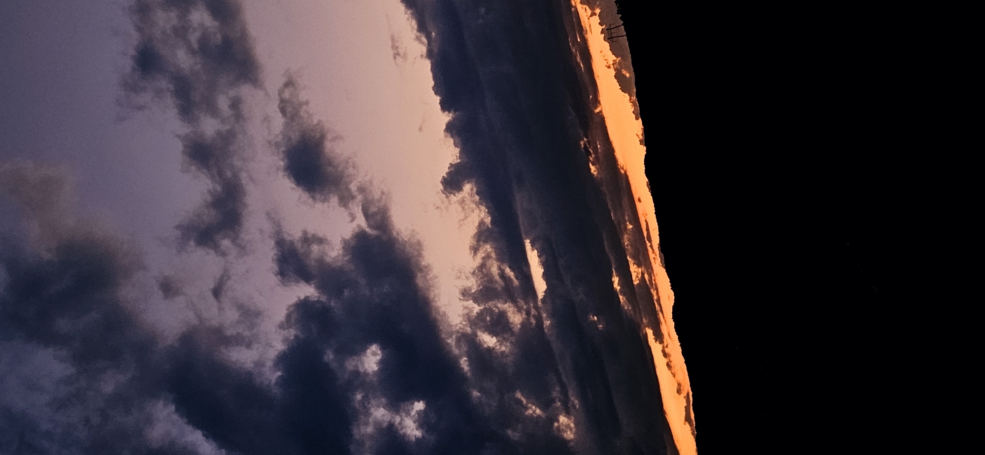 A vertical-orientation photograph captures a sunset or twilight scene over a landscape in Gutsal, Bulgaria. The sky transitions from a deep purplish-blue at the top to a vibrant orange and reddish hue at the horizon. Dark, dense clouds are scattered across the upper two-thirds of the image, appearing as heavy, undulating masses that partially obscure the lighter sky colors. Some clouds along the horizon exhibit orange and pink illumination from the setting sun. A distinct, dark silhouette of a distant landmass, consistent with hills or mountains, runs diagonally across the lower portion of the image, separating the bright horizon from the foreground. A utility pole with cross arms is visible in silhouette against the bright orange sky on the right side of this landmass. The bottom quarter of the image is rendered completely black, providing no discernible detail of the immediate foreground. No human subjects, animals, or specific actions are depicted.