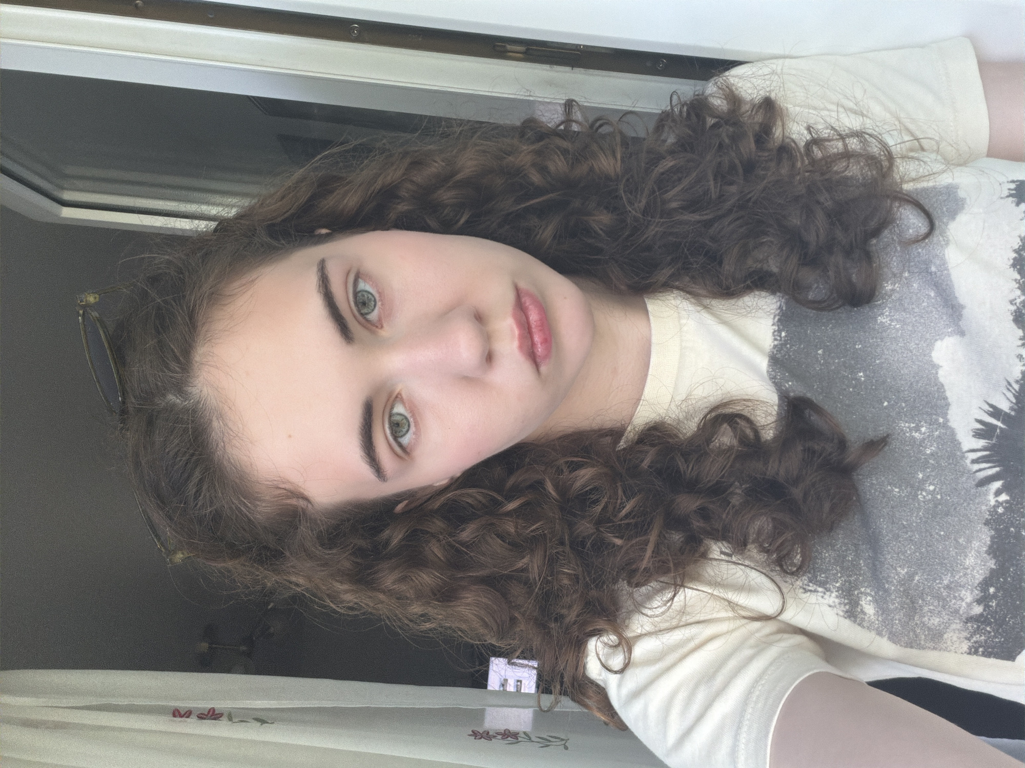 A young woman with curly brown hair and green eyes looks directly at the camera. She is wearing a light-colored t-shirt with a faded graphic design. Her hair is down, falling around her shoulders. She has a neutral expression on her face. The background is blurry, but appears to be an indoor setting with a window and curtains. There are no identifiable landmarks or elements that would suggest a specific location in Zavodoukovsk, Russia.