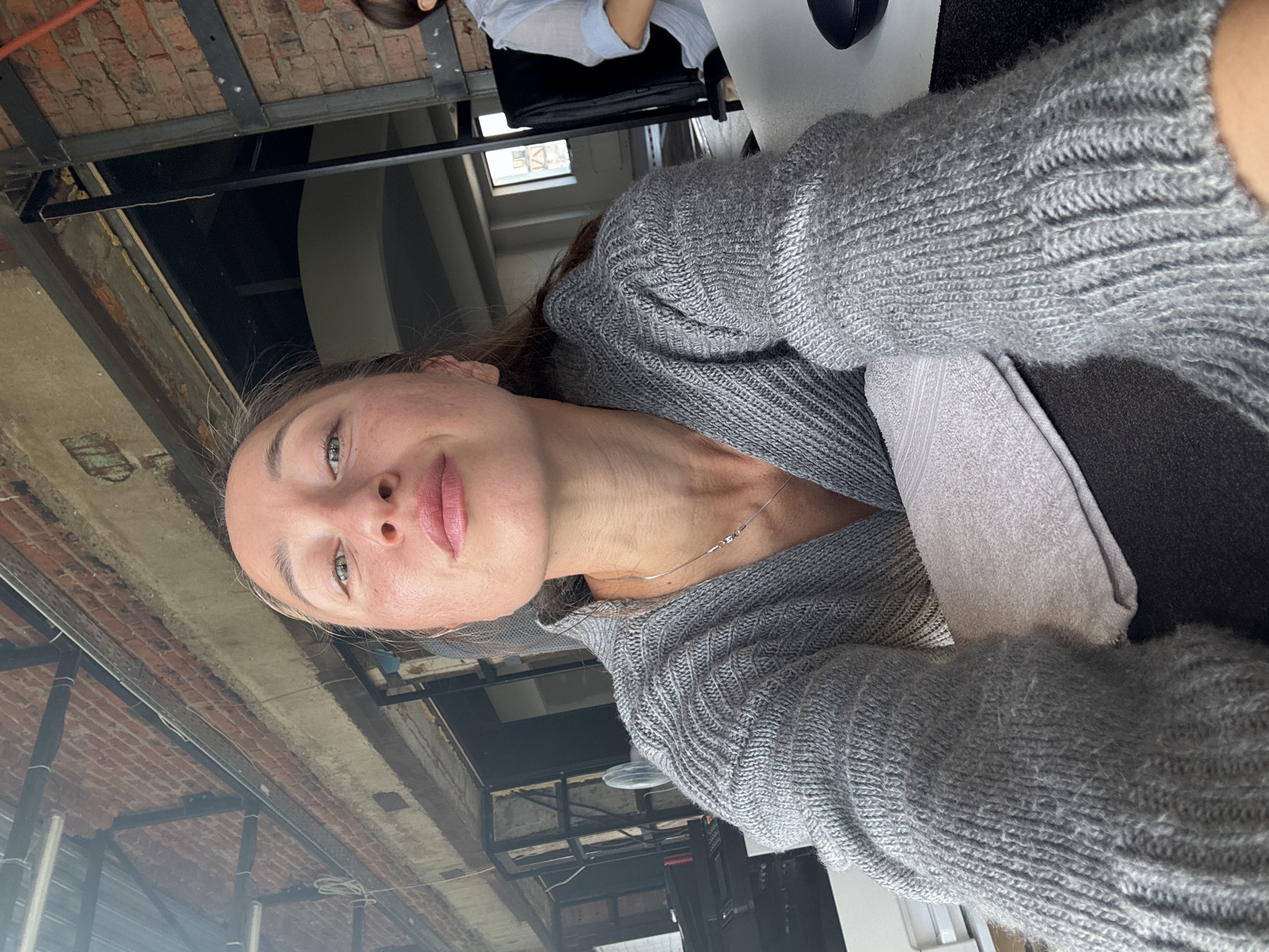 In the foreground of the image, a woman is captured in a close-up selfie. She is wearing a gray knitted sweater, and her head is tilted upwards, looking directly at the camera with a gentle smile. Her makeup appears natural, highlighting her features. A thin silver necklace adorns her neck. A light-colored towel is draped over her arms. In the background, the setting appears to be an indoor space with exposed brick walls and industrial-style ceilings, suggesting a loft or warehouse-style environment. There are hints of office furniture, including desks and chairs, and a window with a view of buildings. The overall impression is a casual, personal moment captured within an urban, possibly repurposed industrial, setting in Chaikovsky, Russia.