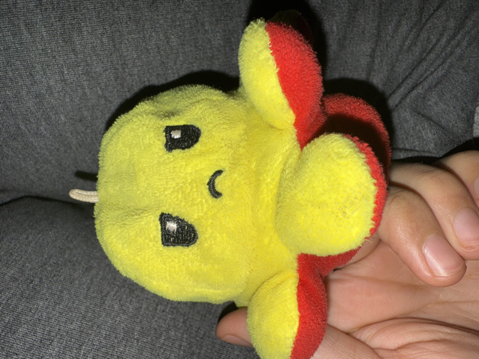 An indoor scene in Bakurtsikhe, Georgia, depicts a close-up of a reversible plush octopus toy held by a human hand. The toy is primarily yellow on its visible side, featuring an embroidered black face with oval eyes and a downturned crescent mouth, indicating an unhappy expression. The underside and tips of its eight tentacles are red. A small, light-colored fabric loop extends from the top of the plush. A human hand, with discernible skin and fingernails, supports the toy from below, with the thumb visible on the lower left. The background consists of a dark grey, textured fabric, likely clothing or upholstery, which is out of focus.