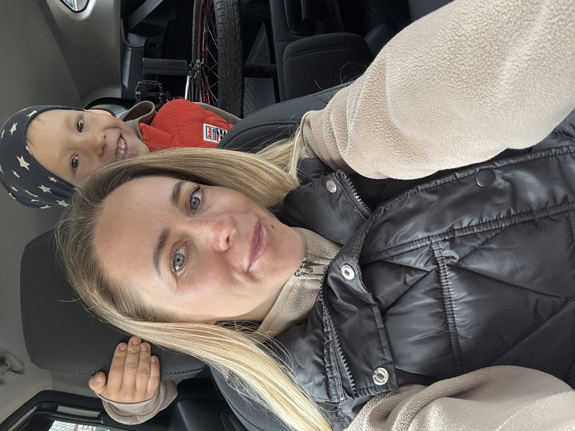 A woman with blonde hair and blue eyes is in a car, taking a selfie. Beside her is a young child wearing a star-patterned hat and an orange vest. A bicycle is visible in the background. The location is Chaikovsky, Russia.