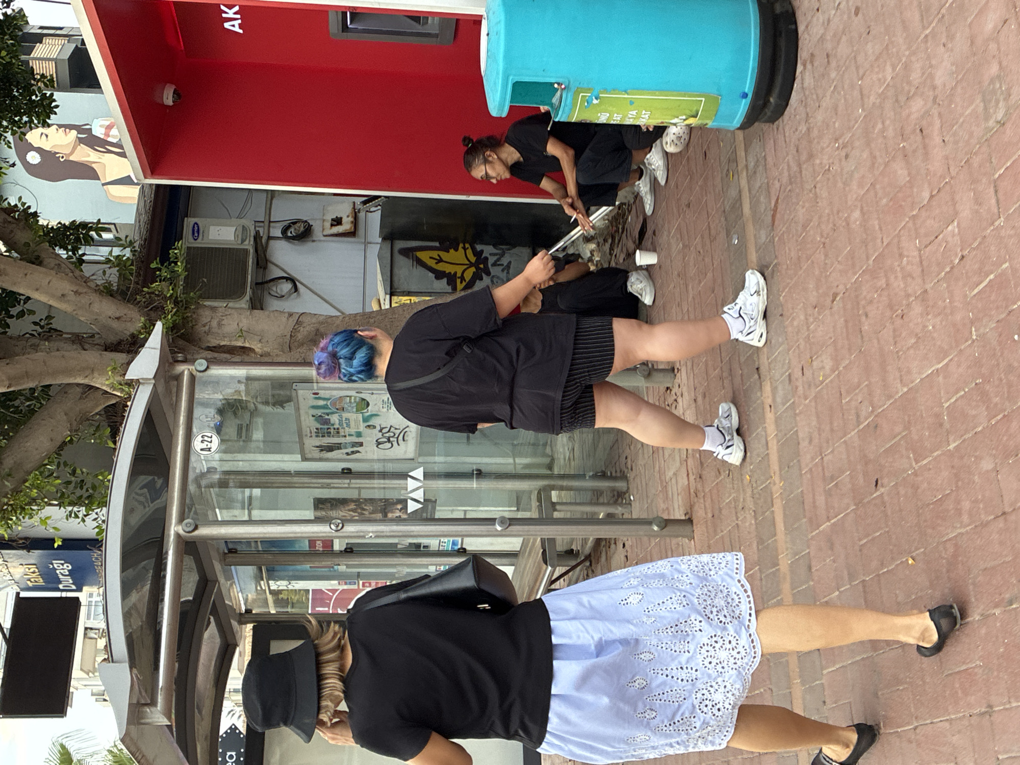 The image captures a street scene in Alanya, Turkey, near a bus stop. In the foreground, a person wearing a black bucket hat and a black t-shirt over a light blue, eyelet-patterned skirt walks away from the camera, facing left. To the right, another individual with vibrant blue and purple dyed hair, wearing a black t-shirt and black and white striped shorts, stands with their back to the camera, looking towards the right.
In the background, near a bright red structure with a security camera mounted on it, two people are seated on the ground. One individual, wearing a black t-shirt and dark pants, is crouched down, appearing to interact with something on the ground or in their lap. Another person, also in dark clothing, is sitting to the right, holding what appears to be a cigarette and looking towards the left. Between them and the red structure is a large, bright blue trash receptacle.
The environment includes a bus stop shelter with glass panels and metal framing. A sign above the shelter reads "TAKSİ DURAĞI" (Taxi Stand). Graffiti is visible on some of the surfaces in the background, including a stylized yellow leaf design on a metal utility box. The ground is paved with red brick-like material. An outdoor air conditioning unit is mounted on a wall behind the bus stop. A mural of a woman's profile is painted on a building in the upper right portion of the image.
