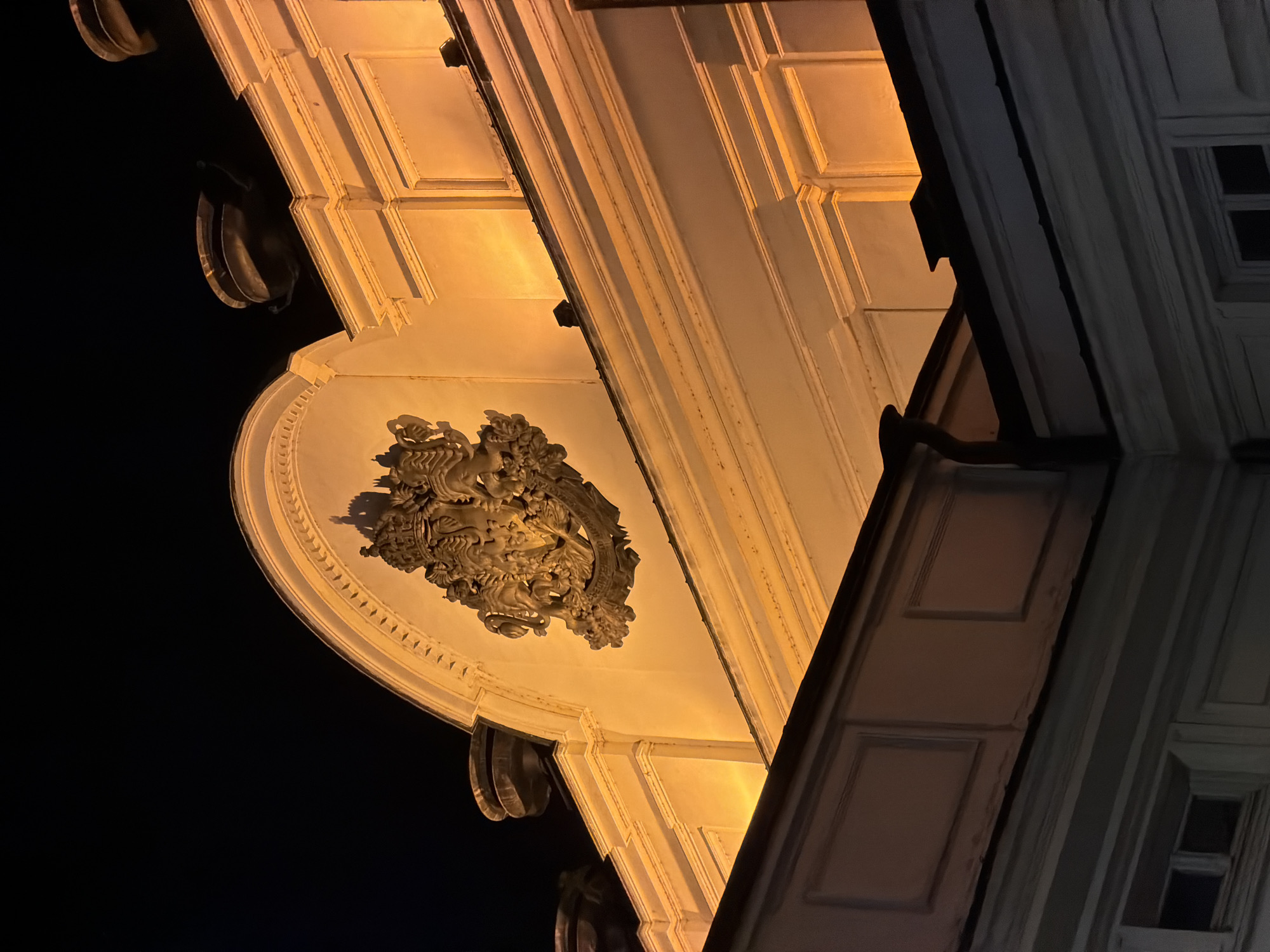 The image shows a close-up, low-angle shot of the façade of a grand building at night, illuminated by warm, golden lights. The architectural style appears classical, with ornate detailing. Prominently featured in the upper center is a large, elaborate coat of arms, intricately carved and detailed, possibly depicting lions, a crown, and other heraldic symbols. The coat of arms is set against a curved pediment adorned with a decorative border of small, pointed elements. Above the pediment, against the dark night sky, are several dark, bowl-shaped decorative elements, possibly urns or finials, on the rooftop.
Below the pediment, the building exhibits layered cornices and recessed panels, all highlighted by the dramatic lighting, creating strong shadows and a sense of depth. In the foreground, a lower section of the building is visible, featuring more rectangular panels and a dark roofline with a drainpipe. The overall impression is one of historical grandeur and solemnity, befitting a significant civic or governmental building. This architectural detail is located on the National Assembly of Bulgaria building in Sofia, Bulgaria.