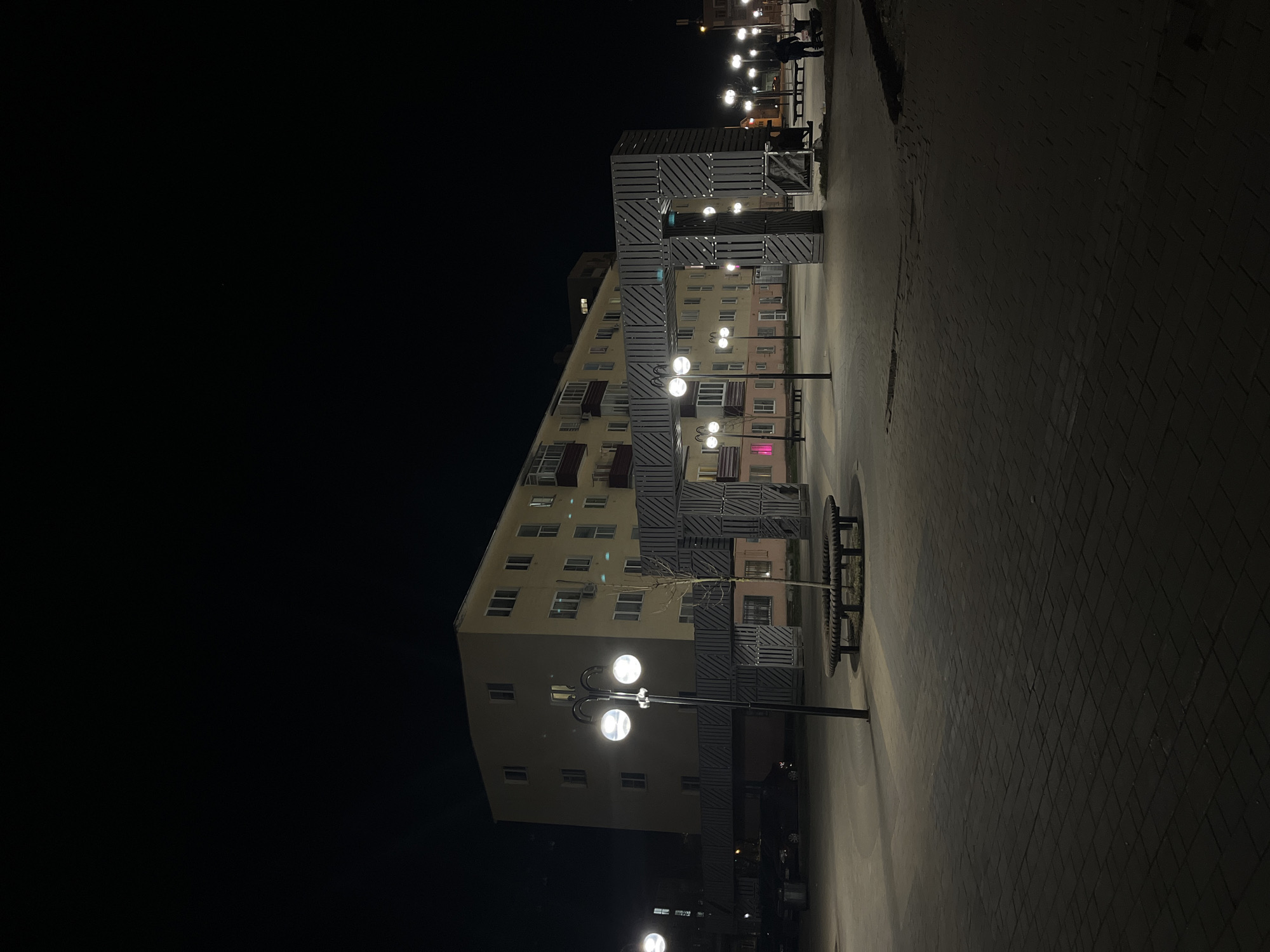 At night in Klin, Russia, a five-story building stands illuminated by street lights. The building is beige with many windows. The façade is decorated with gray, geometric accents. A pathway, lit by globe-shaped street lamps, leads alongside the building. Dimly lit figures are visible further down the path. The area to the left of the building is dark. The ground is paved with dark bricks. A circular bench is situated along the path.
