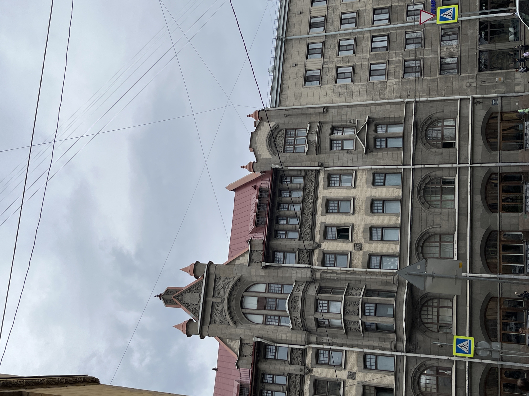 The image shows two adjacent buildings in Saint Petersburg, Russia, under an overcast sky. The buildings are multi-story, with predominantly grey facades. The building on the right is more uniform in design, featuring rows of rectangular windows. The building on the left has more ornate architectural details, including arched windows, decorative moldings, and a red roof. A pedestrian crossing sign is visible in the foreground, indicating a crosswalk. Overhead, power lines intersect the sky.