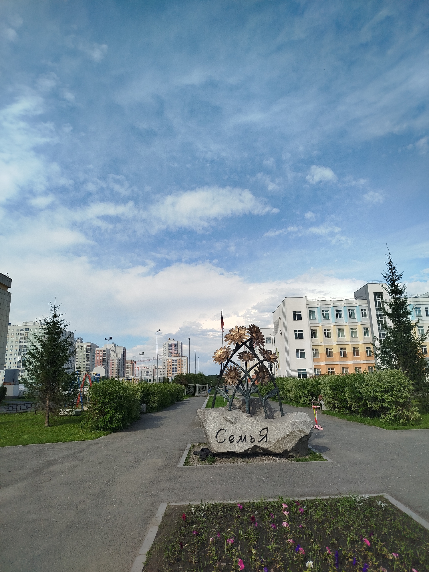 This image depicts an outdoor scene in a residential area, likely a park or public square, with apartment buildings in the background. The primary subjects are a large rock with the Russian word "Семья" (Family) carved into it, and an artistic metal sculpture resembling sunflowers placed on top of the rock. A small pink and yellow scooter is propped against the rock.
The scene is set during the daytime under a partly cloudy sky. The weather appears pleasant, with the sun casting light and shadows. The architecture of the buildings suggests a modern urban environment. Green grass, manicured bushes, and a flowerbed with colorful flowers are visible in the foreground, indicating a well-maintained public space. A tall evergreen tree stands to the left of the sculpture.
There are no people visible in the frame, and no specific activity or event is taking place, suggesting a peaceful, everyday setting. The overall mood is serene and perhaps slightly nostalgic due to the word "Family" and the floral motifs. The location cues, such as the apartment buildings and the Cyrillic text, combined with the provided location context of Yekaterinburg, Russia, help identify the setting.