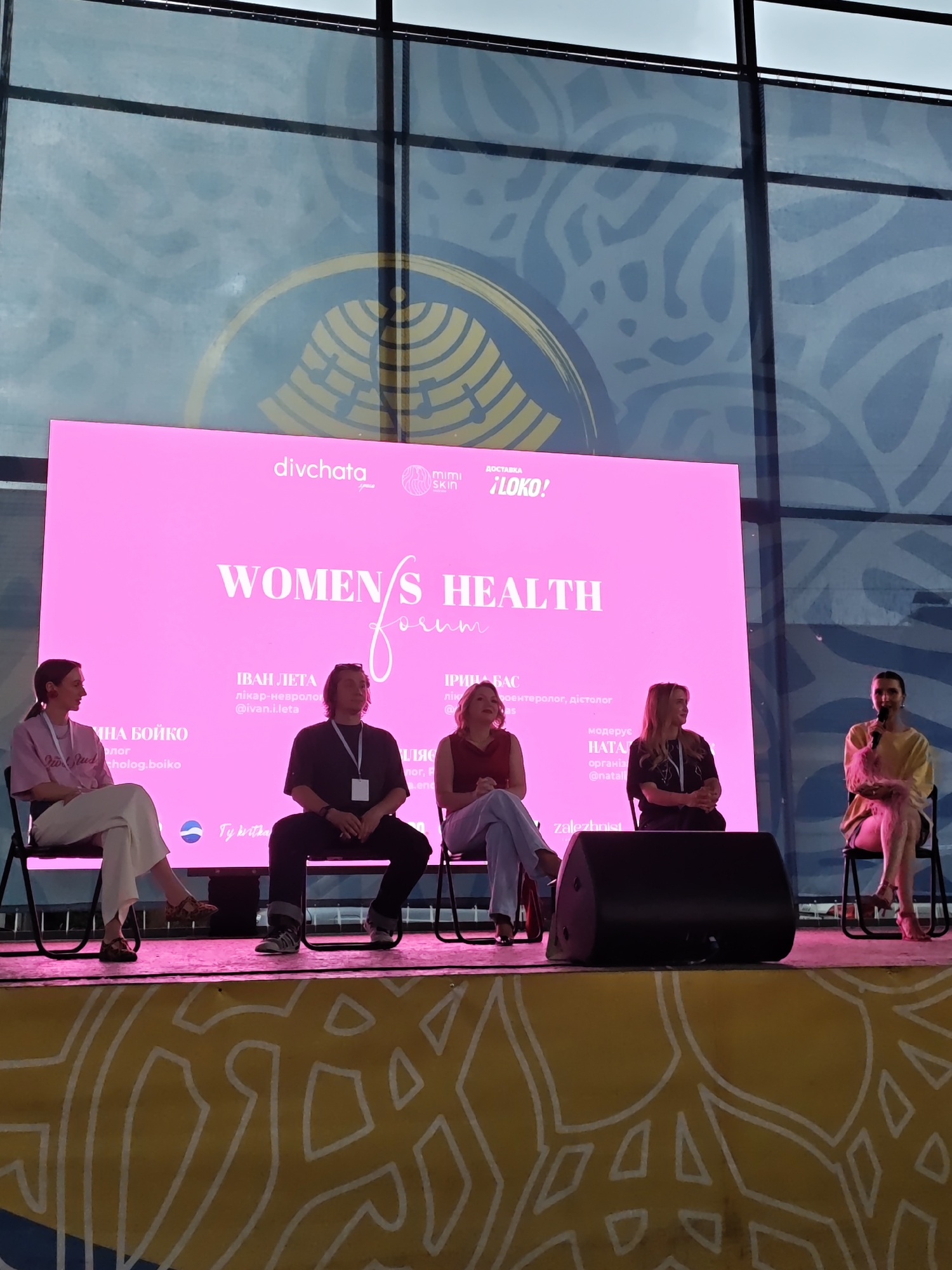 The image depicts a panel discussion taking place in Vinnytsia, Ukraine, featuring five individuals seated on a stage against a large pink LED screen. The background behind the screen consists of large multi-pane windows, showing a bright, slightly overcast sky and a faint, abstract, light blue pattern with a partial golden circular motif. On the prominent pink screen, the main title "WOMEN'S HEALTH forum" is displayed in white text, with "forum" in a cursive script. Above the title, logos for "divchata" and "mimi skin" are visible on the left and right, respectively, along with "ДОСТАВКА !LOKO!". Below the main title, speaker names, roles, and social media handles are listed: "ІННА БОЙКО психолог", "ІВАН ЛЕТА лікар-невролог", and "ІРИНА БАС лікар-гастроентеролог, дієтолог". Further to the right, "модерус НАТАЛІЯ" is partially visible. At the bottom of the screen, a small logo of a blue sphere with a white wave and the text "Ty Kvitne" are present. From left to right on the stage, the individuals are: 1. A woman with dark hair in a light pink t-shirt and off-white wide-leg trousers, wearing dark patterned sandals. She sits with her hands clasped in her lap. The screen identifies her as "Інна Бойко, психолог." 2. A man with shoulder-length brown hair, wearing a dark short-sleeved shirt, dark trousers, and athletic shoes. He has an ID badge on a lanyard and sits with hands clasped. The screen identifies him as "Іван Лета, лікар-невролог." 3. A woman with light hair, wearing a maroon short-sleeved top and light blue trousers, with dark high heels. She sits with hands clasped, looking towards the right. The screen identifies her as "Ірина Бас, лікар-гастроентеролог, дієтолог." 4. A woman with dark hair, wearing a black top with white detailing and dark trousers. She sits with hands clasped. 5. A woman with dark hair, wearing a light yellow long-sleeved top and light shorts or skirt. She holds a microphone with a pink feathery accessory and is wearing pink high heels. Her legs are crossed, and she appears to be speaking. The screen identifies her as "Наталія" (moderator). All five individuals are seated on black folding chairs. On the stage floor in front of the man, a large black stage monitor or speaker is visible. The stage itself is covered with a yellow fabric or banner featuring a repeating white abstract geometric pattern. The lighting on the stage is bright, with a noticeable pink