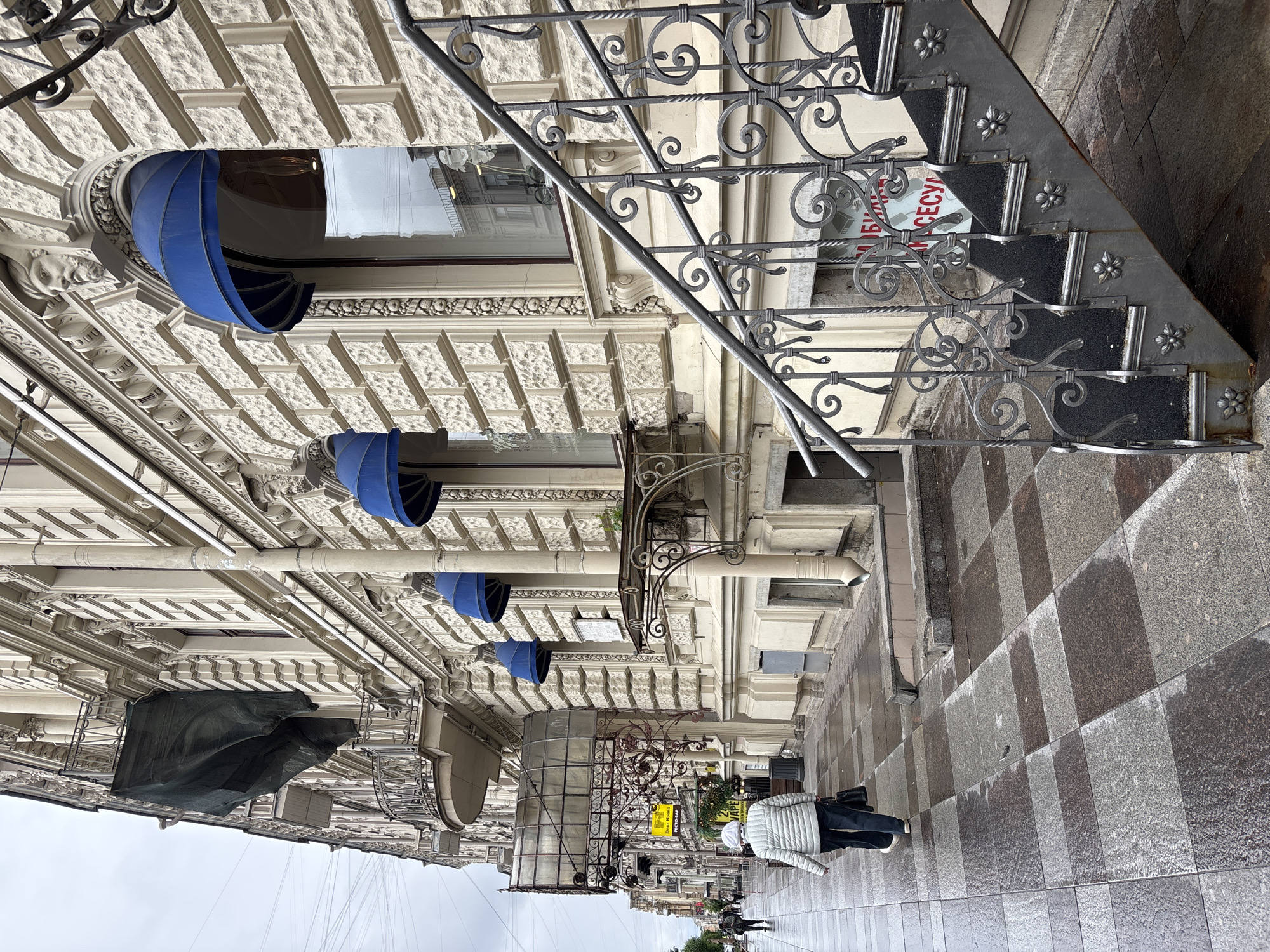 The image depicts a street scene in Saint Petersburg, Russia, characterized by its ornate architecture and wet pavement, indicating recent rain. The perspective suggests the photo was taken at ground level, looking up at the buildings.
**Architecture**: The buildings are adorned with elaborate facades, featuring intricate details, stucco work, and blue awnings over the windows. One building is partially covered in scaffolding and a black tarp, indicating ongoing construction or renovation work. The buildings' style is typical of historic architecture in Saint Petersburg.
**Street Level**: The sidewalk is paved with alternating light and dark grey square tiles. The pavement is wet, reflecting the building facades.
**People**: Several individuals are walking along the sidewalk. One person is wearing a light-colored jacket, dark pants, and a white head covering, walking towards the right. Other people are walking in the background, further down the street.
**Objects**: A black wrought-iron staircase is prominent on one of the buildings. It has ornate detailing and small floral embellishments. A sign in Russian is visible near the staircase. Various pipes and architectural fixtures are attached to the buildings.