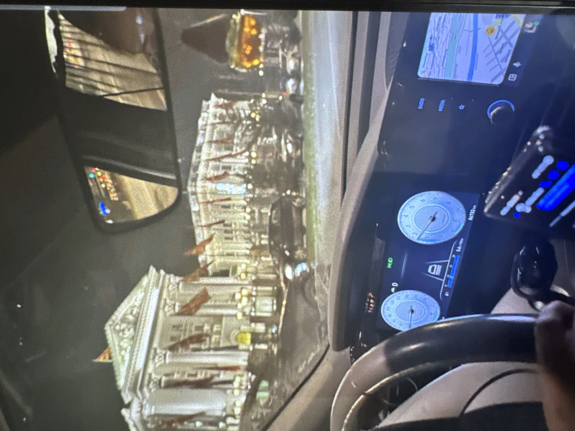 A nocturnal image captured from inside a vehicle presents a detailed view of a city street and architectural structures in Vratsa, Bulgaria, through the windshield and a partial view of the vehicle's interior.
The external scene, viewed through the windshield, features two prominent, grand classical-style buildings illuminated brightly against a dark sky. The building on the left exhibits a triangular pediment atop a facade with columns, displaying multiple stories and windows. Both buildings are adorned with numerous red or orange rectangular flags or banners, evenly spaced across their fronts. In front of these structures, a street is visible with several vehicles, including sedans and SUVs, parked or in motion. Interspersed between the street and the buildings are trees or shrubs, some appearing to be decorated with lights. Streetlights also contribute to the scene's illumination. The upper right portion of the image shows a rearview mirror reflecting blurred lights and vertical elements, possibly from the car's interior or ambient street lights.
The internal scene of the vehicle occupies the lower half of the image. The steering wheel, partially visible on the left, features a dark and a lighter section. The digital dashboard displays two circular gauges with blue backlighting. The left gauge indicates "0 D" with other digital information, while the odometer below it reads "86732 km." The right gauge shows an RPM or speed reading. To the right of the instrument cluster, a larger infotainment screen displays a navigation map with a blue and gray color scheme. Text labels "RADIO" and "MEDIA" are visible above physical buttons on the right side of the screen. Below the primary screens, additional vehicle controls and illuminated blue elements are present. A human hand is partially visible in the bottom-left corner, positioned near the steering wheel.