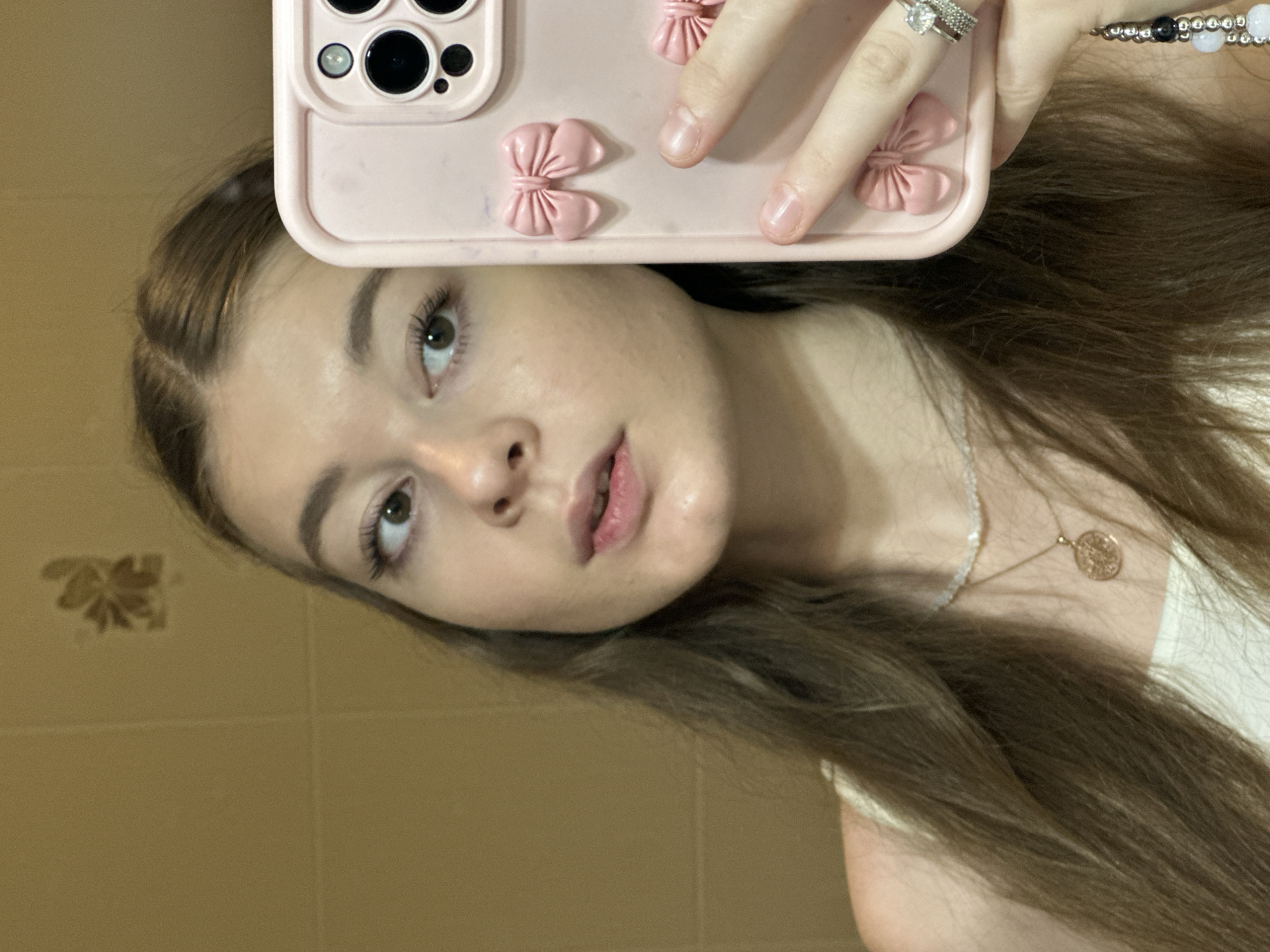 The image shows a young woman taking a mirror selfie. She has long, brown hair and is wearing a white top. She is also wearing a necklace with a pendant and rings on her fingers. The background appears to be a tiled wall, possibly in a bathroom. The woman is holding a pink phone case with bows on it. There is no indication of the location being Kazan, Russia, nor any specific details about the city.