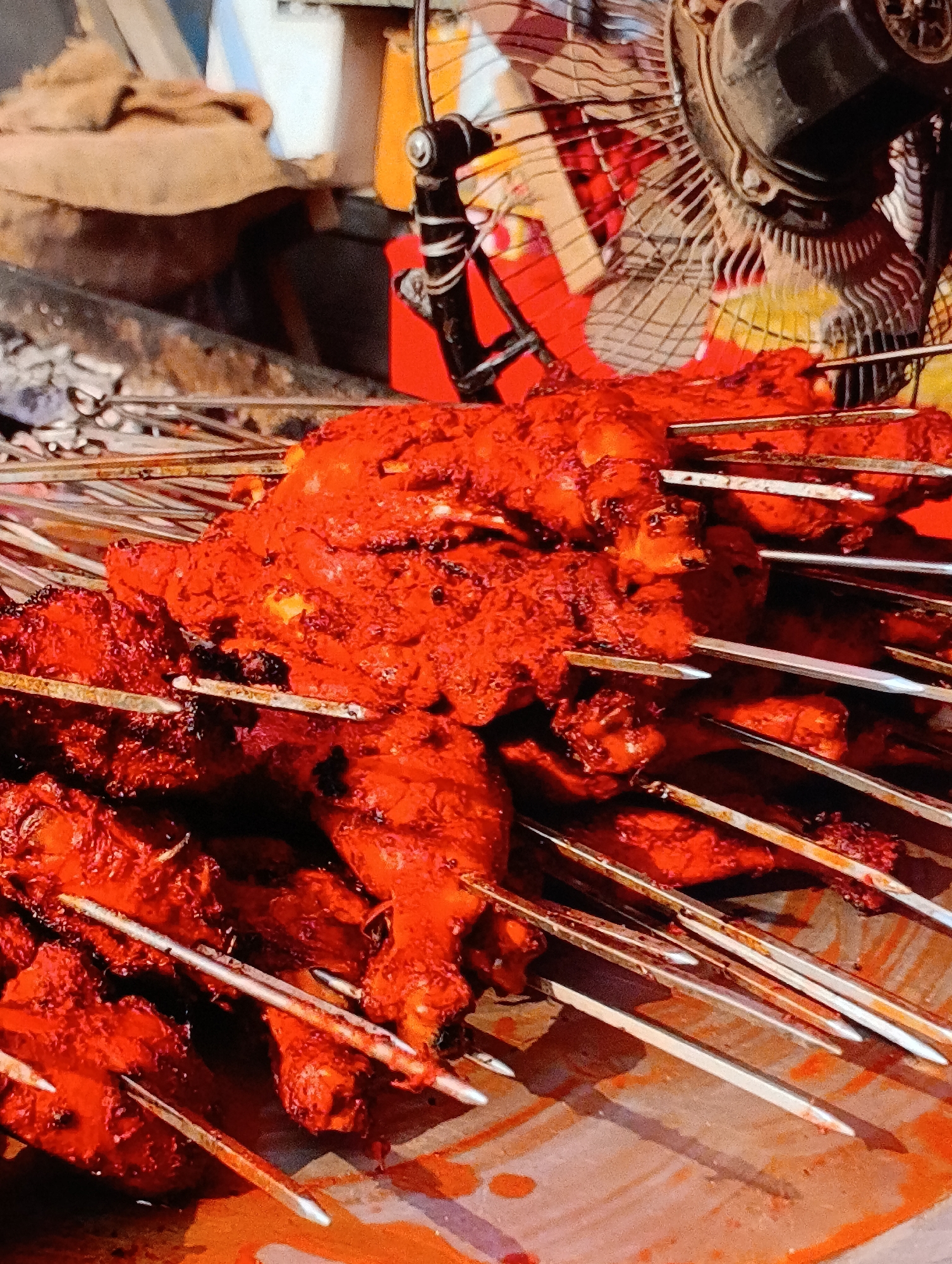 The image depicts a close-up of skewered, red-marinated chicken pieces stacked on a surface. Skewers protrude from the meat at various angles. In the background, an older-style fan is visible, along with a glimpse of what appears to be a cooking area with sacks and potentially embers. The location is identified as Unknown, Unknown.