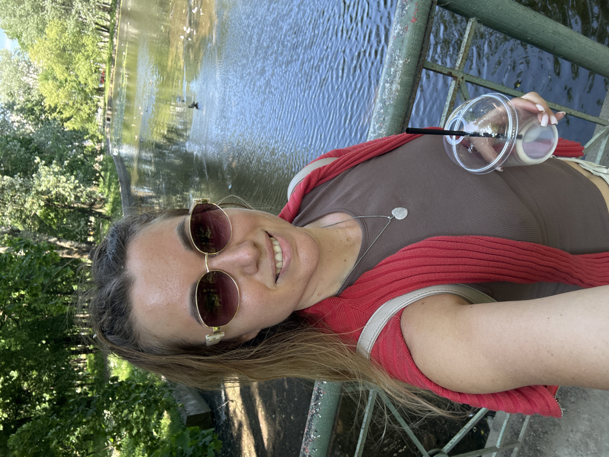 A woman in sunglasses takes a selfie by a body of water in a park. She is wearing a brown tank top, a red sweater draped over her shoulders, and a silver pendant necklace. She holds a clear plastic cup with a black straw in her right hand. The water in the background has several birds swimming in it. Trees and foliage surround the water. A green railing is visible in the foreground.
