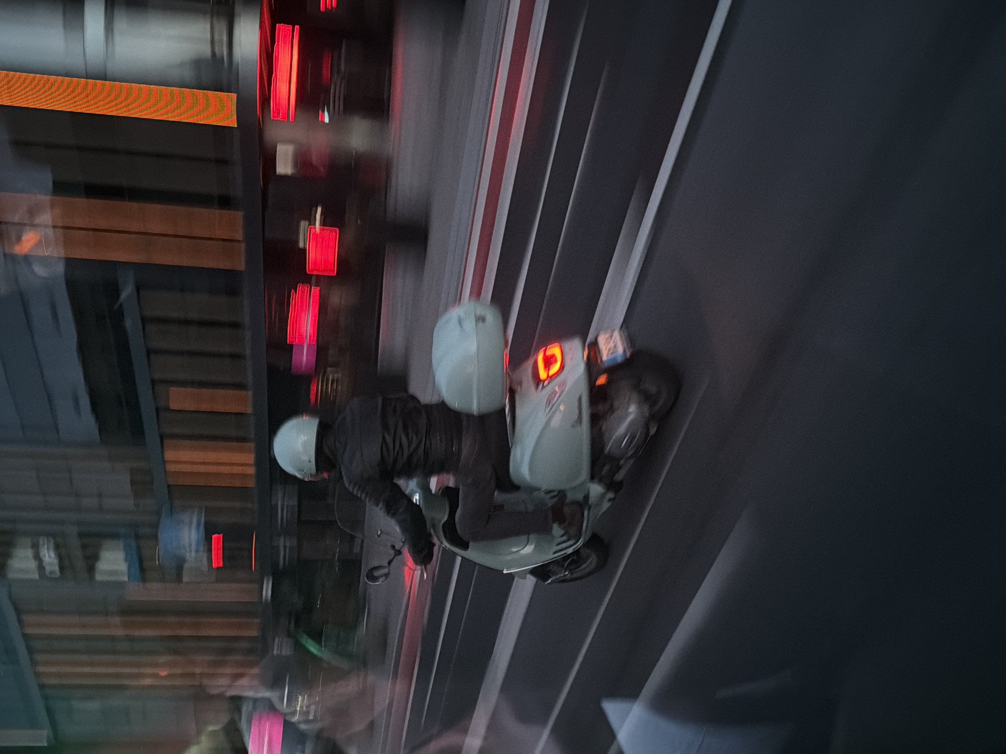 A person wearing a light blue helmet and a black leather jacket rides a light blue scooter in the city of Tallinn, Estonia. The scooter has a light blue storage box on the back. The scooter is on a road with white and red markings. In the background, there are buildings with vertical orange and brown panels and red lights. The image is blurred due to motion.