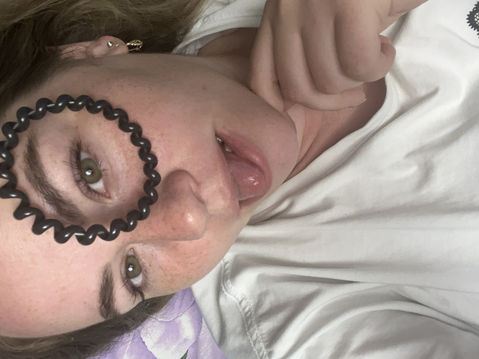 A woman with light skin is lying down. She has green eyes and freckles. A black coiled hair tie partially covers her left eye. She is sticking her tongue out. Her right hand is near her neck. She is wearing a white t-shirt. There is a hint of purple fabric in the lower left corner. The location and city of Chaikovsky, Russia, cannot be determined from the image.