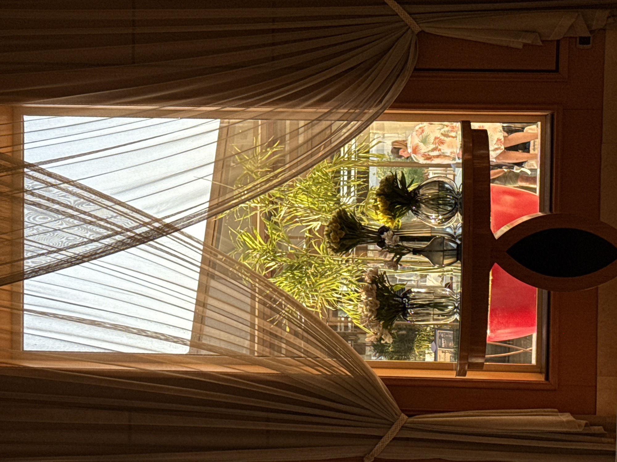 The image depicts an indoor scene looking out through a large window with sheer, crisscrossed curtains. On a polished wooden table in the foreground, several glass vases hold floral arrangements. The background reveals a sunlit outdoor area with lush green foliage, possibly bamboo. Through the window, a person in a floral shirt and white skirt is partially visible, standing near a red object. The setting appears to be a hotel or resort in Sharm El Sheikh, Egypt, suggested by the tropical plants and the overall ambiance.