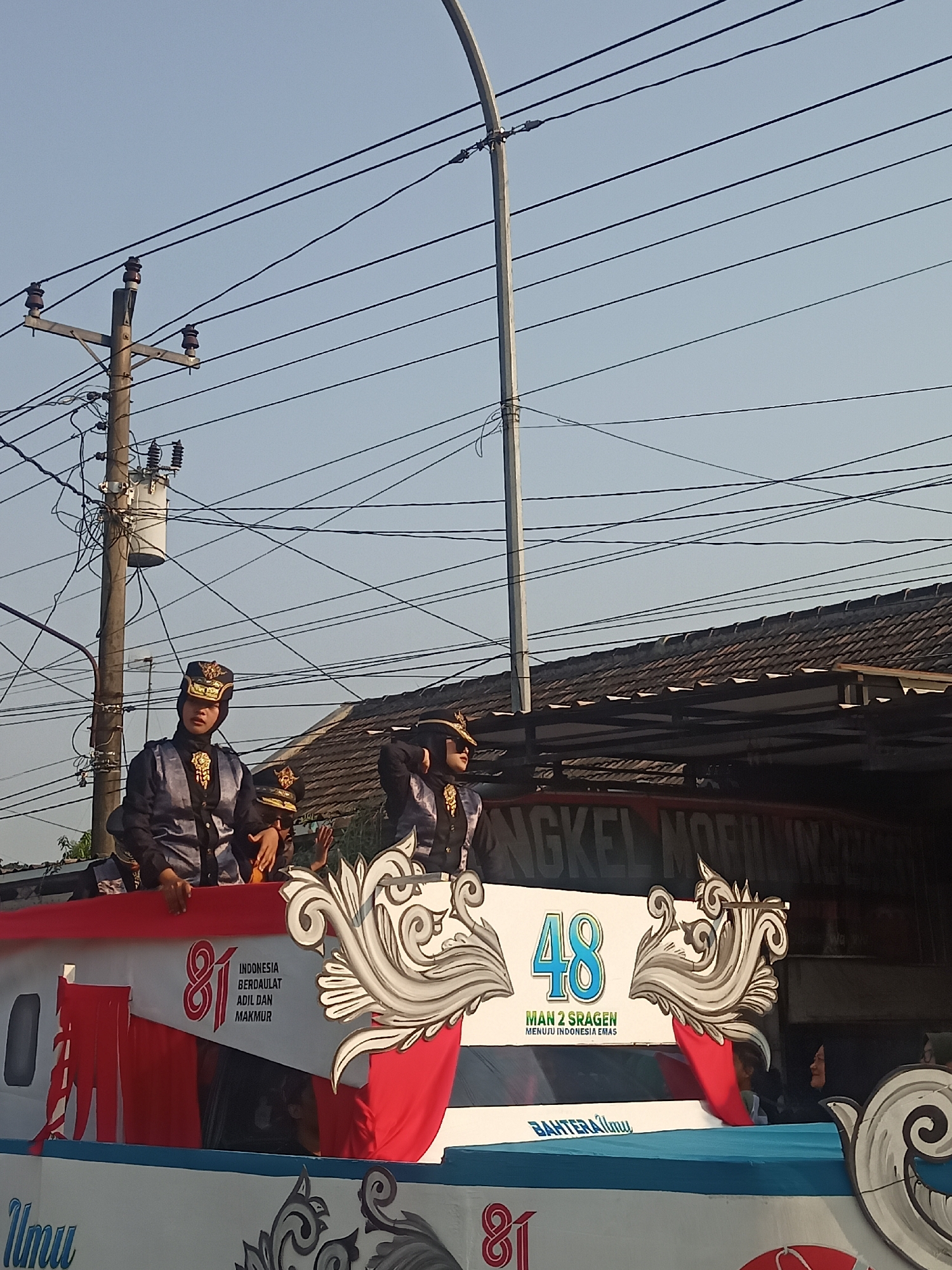 The image shows a parade float with several individuals in traditional attire. The float is decorated with red and white elements, and features text that includes "INDONESIA BERDAULAT ADIL DAN MAKMUR" and "48 MAN 2 SRAGEN MENUJU INDONESIA EMAS". The individuals are wearing dark uniforms with gold accents and ornate headwear. In the background, there are buildings with tiled roofs and utility poles with numerous electrical wires. The scene appears to be outdoors on a sunny day, likely on a street in Ngrompongan, Indonesia, given the text on the float.