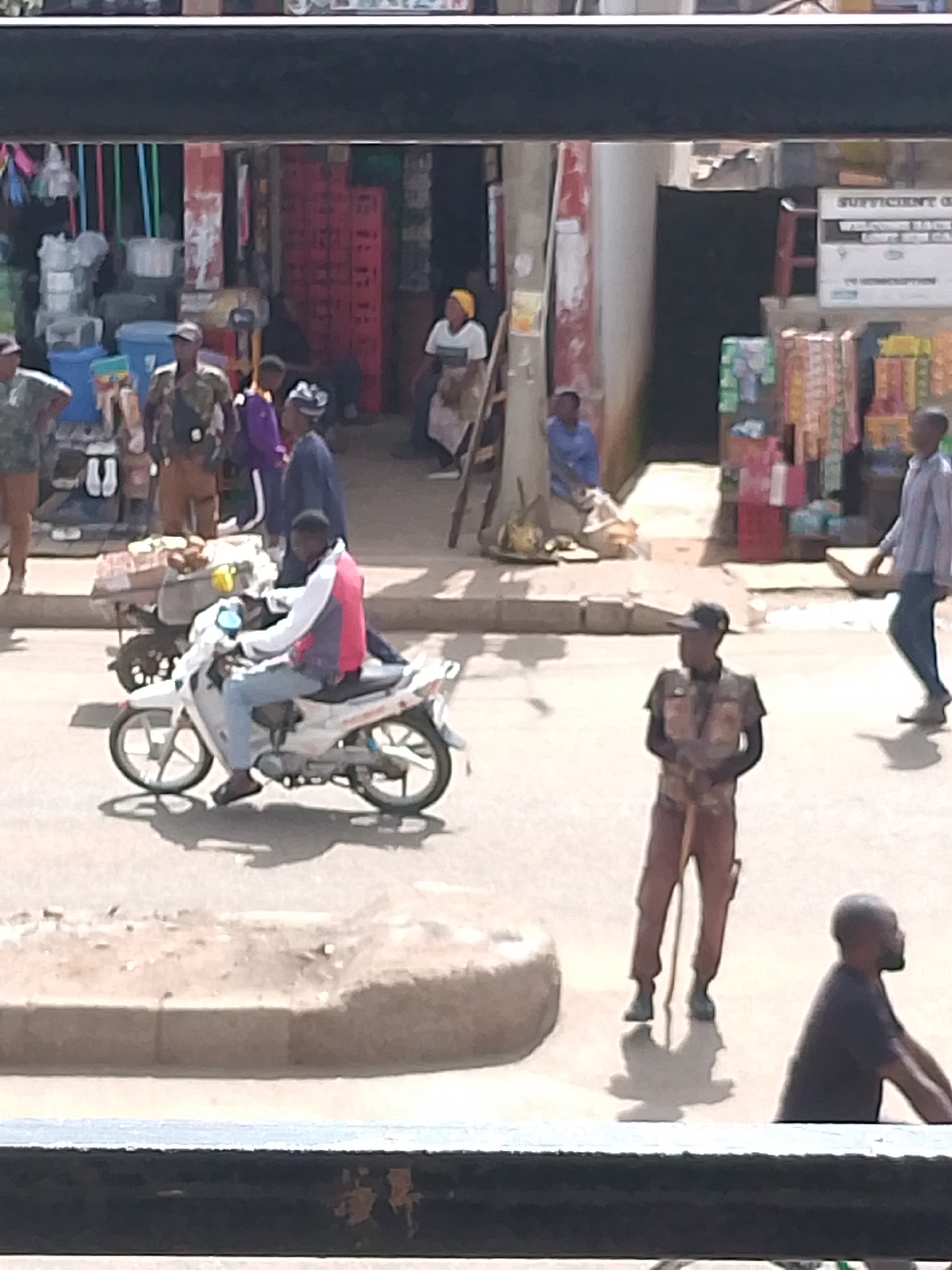 The image depicts a street scene in Bukuru, Nigeria, with several individuals engaged in various activities. In the foreground, a person is riding a white motorcycle with a multi-colored jacket. Behind the motorcycle, a uniformed individual stands with a rifle. Further in the background, multiple people are seen near market stalls. Some are browsing, while others appear to be vendors. The environment suggests a bustling marketplace with an assortment of goods displayed for sale. The architecture suggests a typical urban setting in Nigeria, with market stalls lining the street and people going about their daily routines.