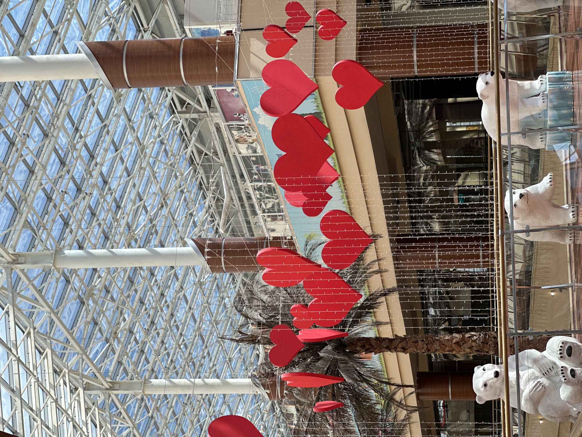 The image shows an indoor atrium decorated with numerous red heart-shaped ornaments and strings of white lights. In the foreground, there are three white polar bear sculptures, two of which are fully visible. The bears are positioned on a platform with a railing. Behind the sculptures, a palm tree is visible, adorned with some of the red hearts. The ceiling of the atrium is made of glass and metal framework, allowing natural light to filter in. The background shows multiple levels of balconies and architectural elements, suggesting a large commercial or public space. There are no people clearly visible in the image. The location of Dubovoye, Russia, is not evident from the visual information provided in the image.