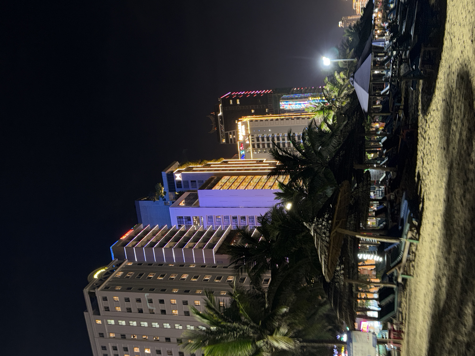 Night scene in Đà Nẵng, Vietnam, showing illuminated high-rise buildings and palm trees. Beach furniture is on the sand.