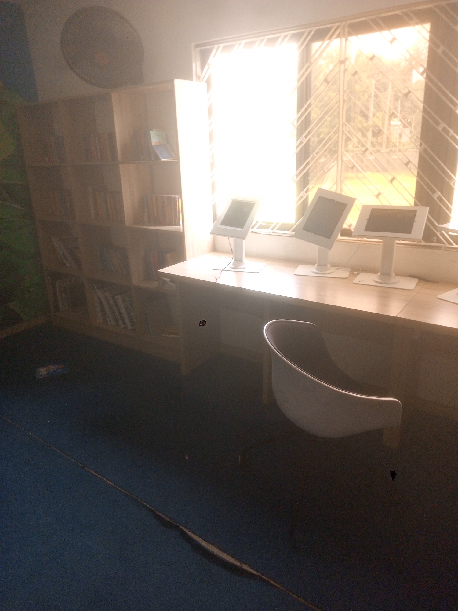 The image shows an interior setting, likely a library or study area in Abuja, Nigeria. A bookshelf filled with books stands against a wall, next to a window with a security grill. Three computer monitors are arranged on a desk in front of the window. A white chair sits in front of the desk. The room is dimly lit, with the light source appearing to be primarily from the window. The floor is blue, and a dark, elongated object is visible on the floor.